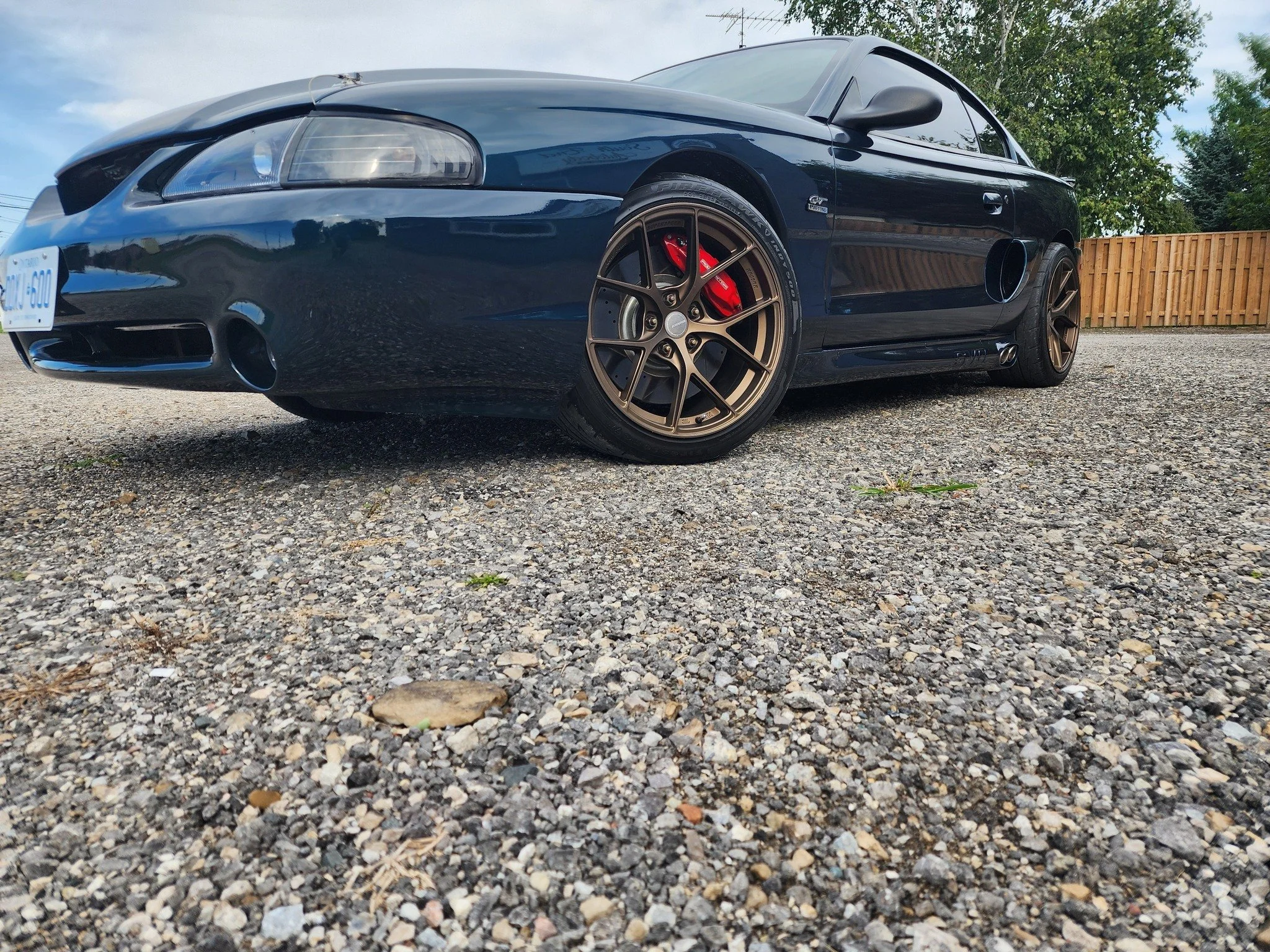 Black sports car with bronze wheels parked on gravel surface, red brake calipers visible through the wheels, and a wooden fence in the background.
