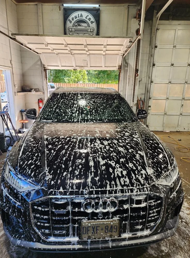 Black Audi car covered in soap suds inside a garage with an open garage door, with a South End sign hanging above.