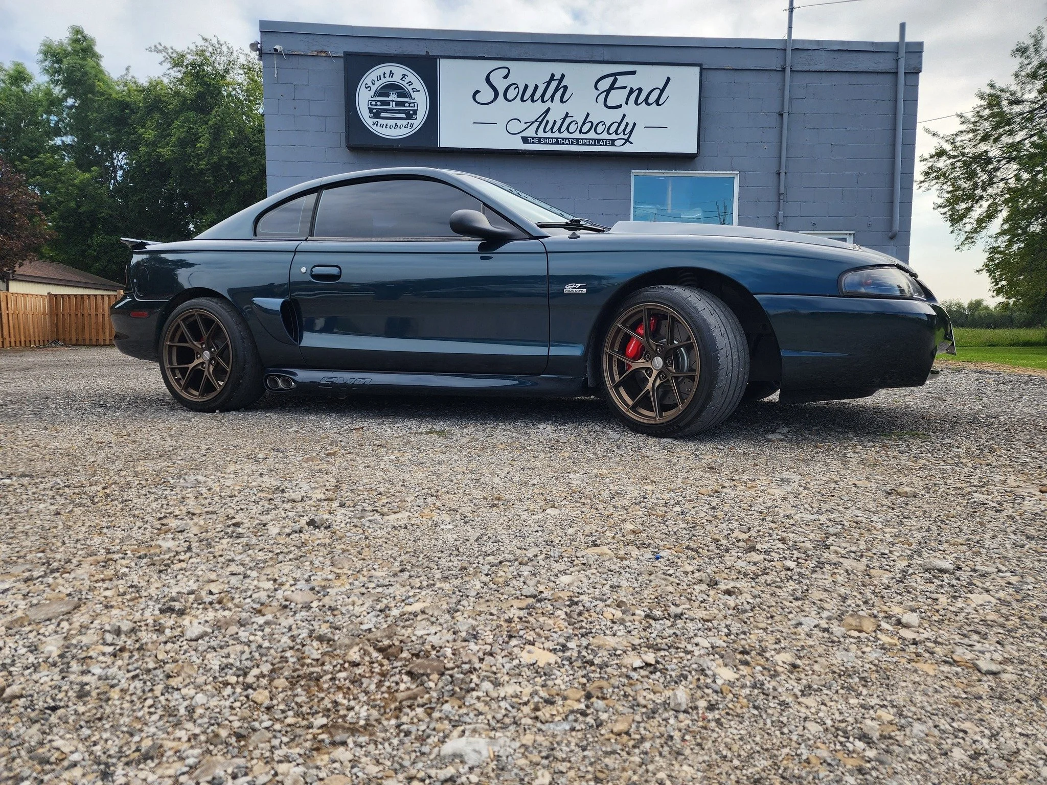 A black sports car parked in front of a gray auto body shop with a sign that reads "South End Autobody." The car has bronze wheels, red brake calipers, and a sleek design, parked on a gravel lot with trees and a fence in the background.