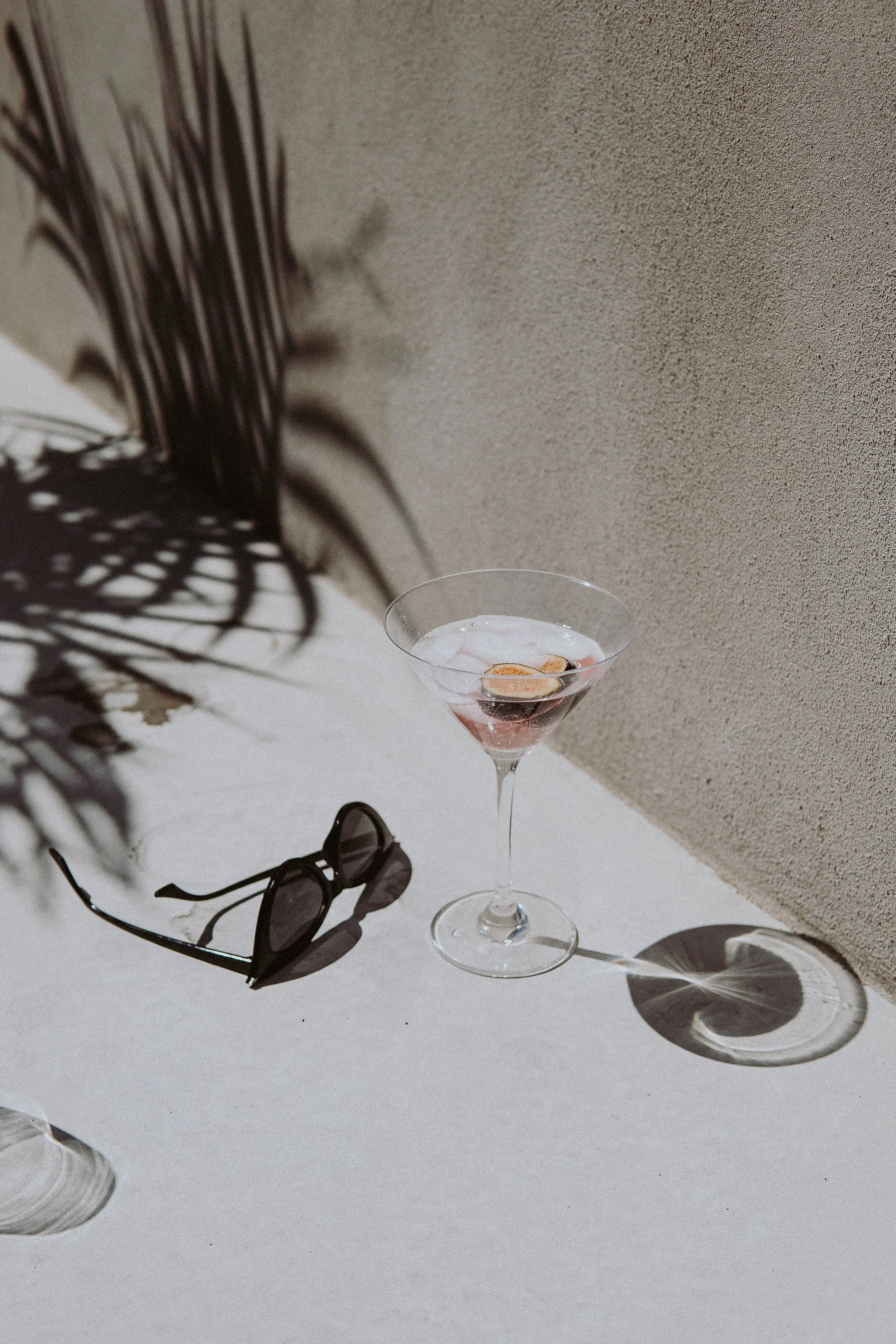 A cocktail in a martini glass with a cherry garnish. A pair of sunglasses lies on the surface next to the glass. Shadows of plants are cast on the surface, with a beige wall in the background.