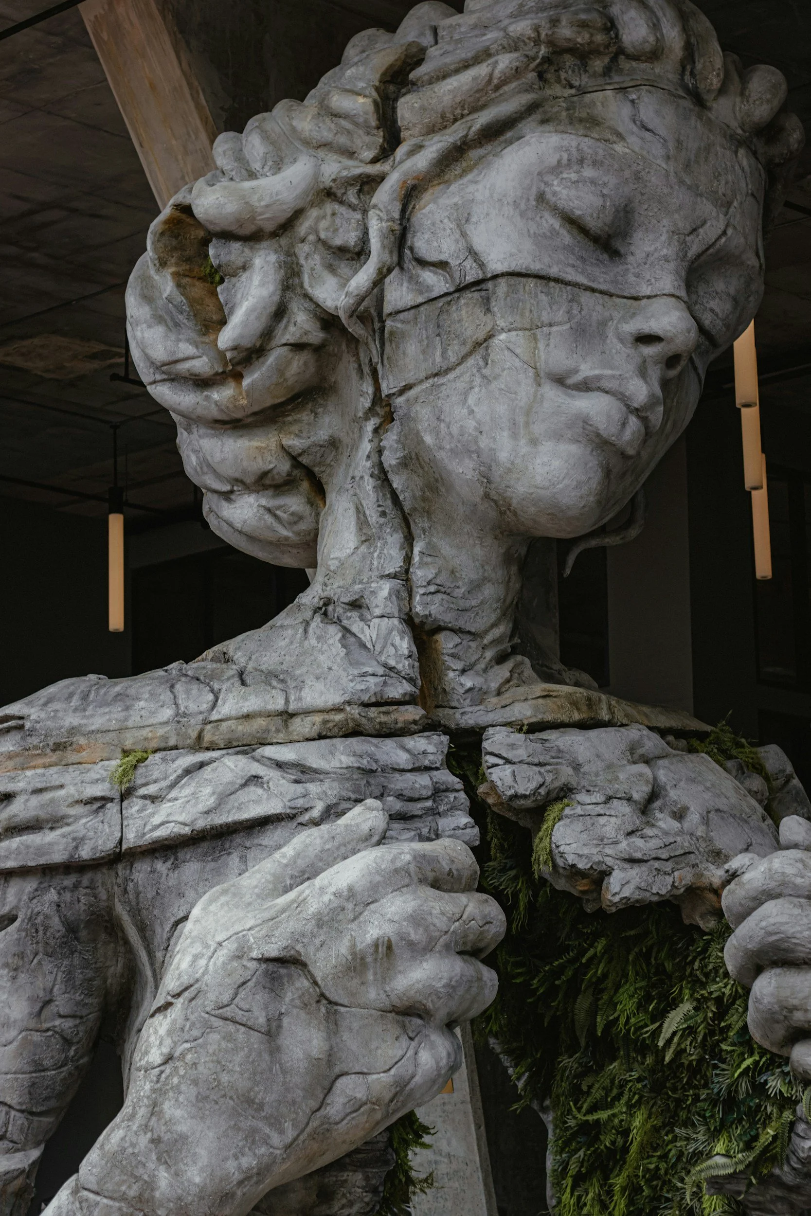 A large stone sculpture of a woman’s face and hands, with her hands holding a tear drop shape, located indoors with modern lighting and decor.