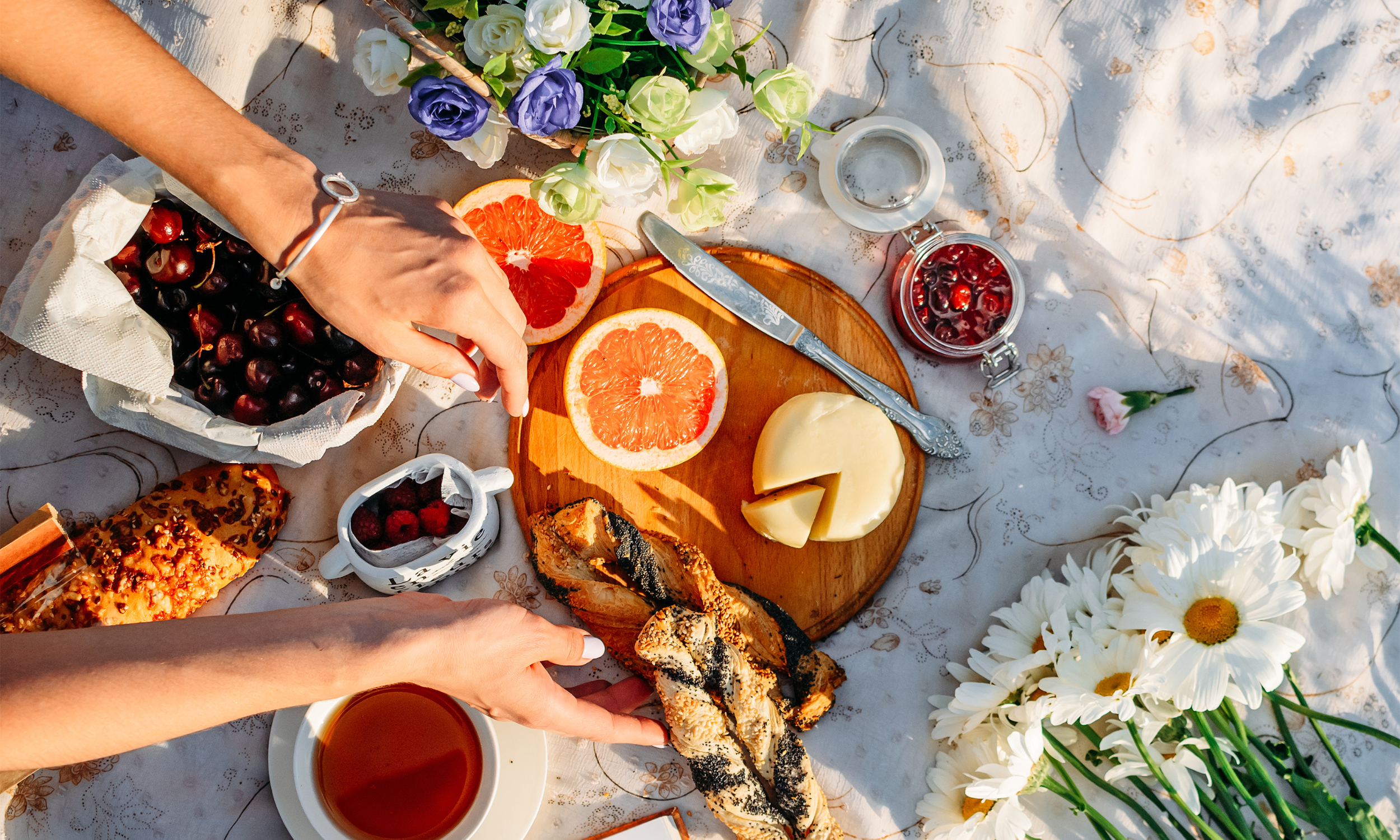 A picnic table with sliced grapefruit, cheese, bread, cherries, raspberries, tea, and flowers on a white embroidered tablecloth.