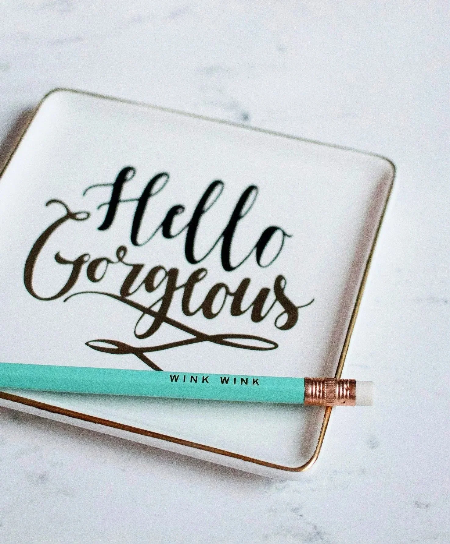 A decorative plate with the words 'hello gorgeous' in black cursive writing on it. A mint green pen with a rose gold tip and the words 'WINK WINK' rests on the plate. The background is a white marble surface.