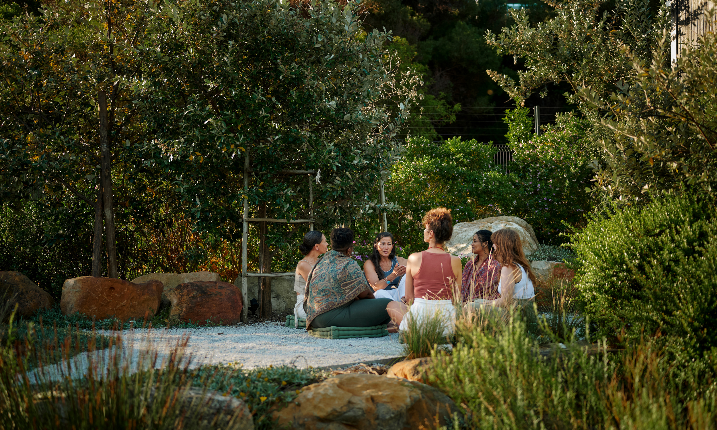 A group of six women sitting on cushions in a circle outdoors, engaging in conversation amidst lush green trees and bushes during daylight.