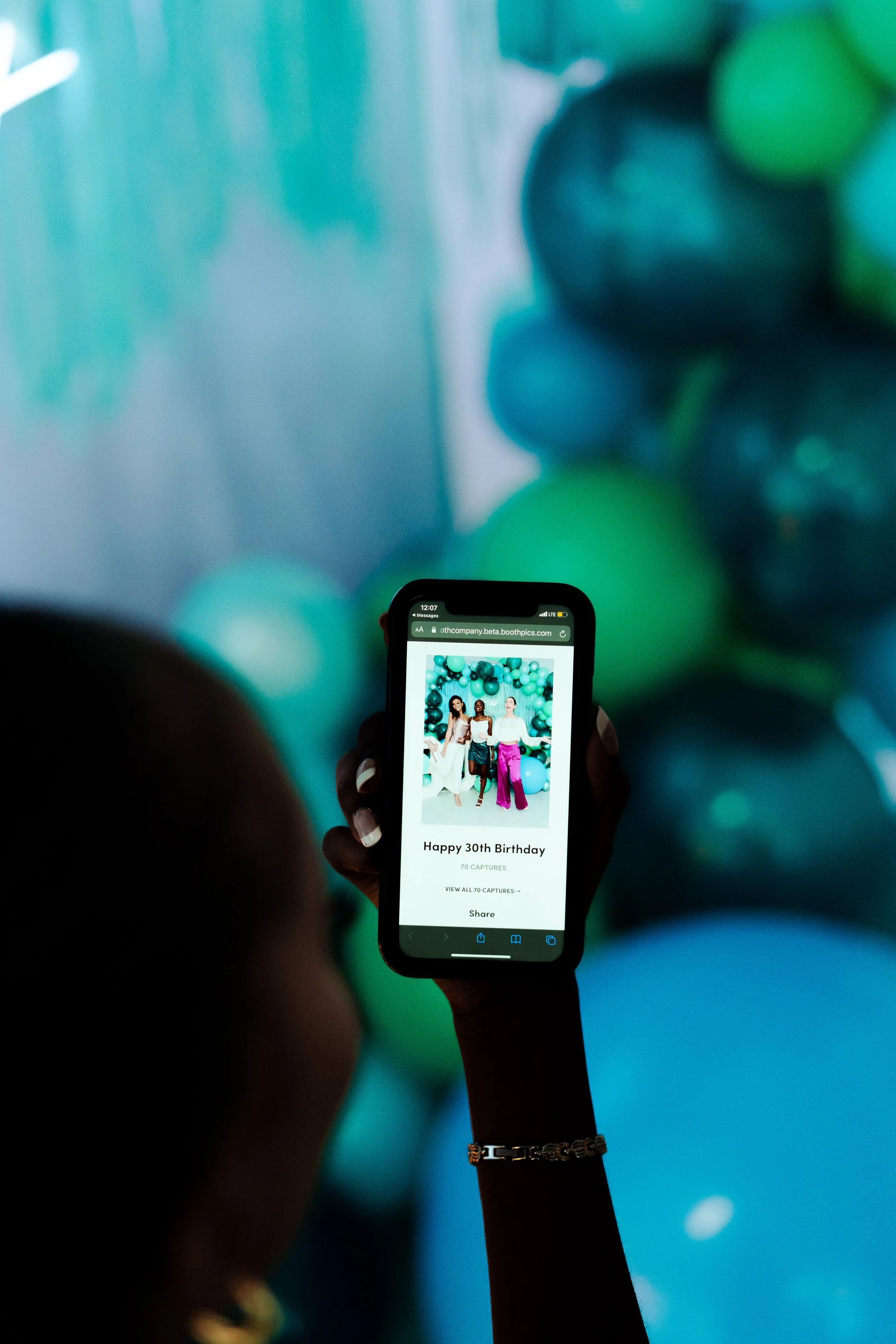 Person holding a phone displaying a birthday celebration photo with three women in front of blue and green balloons. Text on phone reads 'Happy 30th Birthday'.