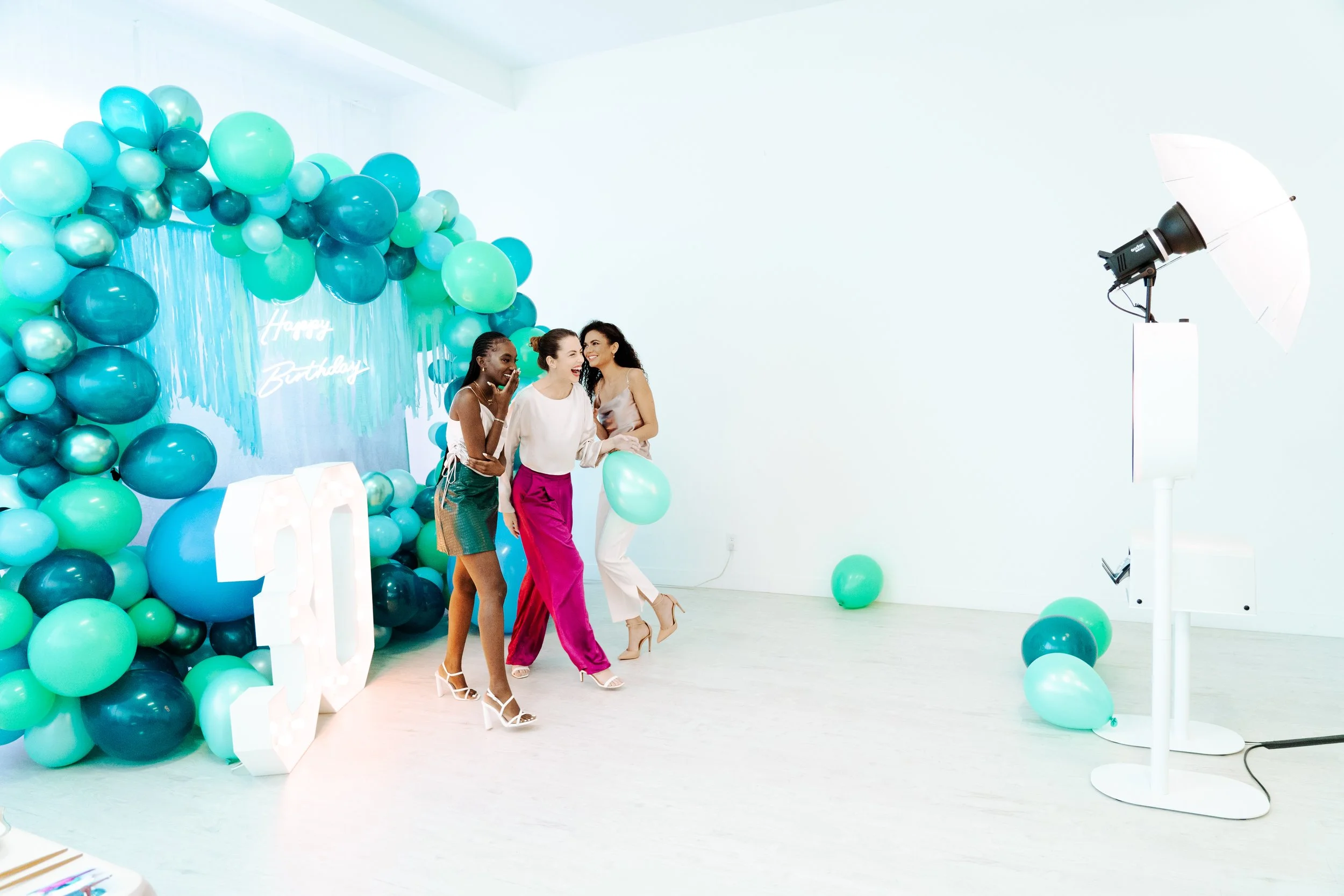 Three women at a birthday celebration, laughing and posing in front of a balloon arch with a 'Happy Birthday' sign, colorful balloons, and a photo backdrop with studio lighting.