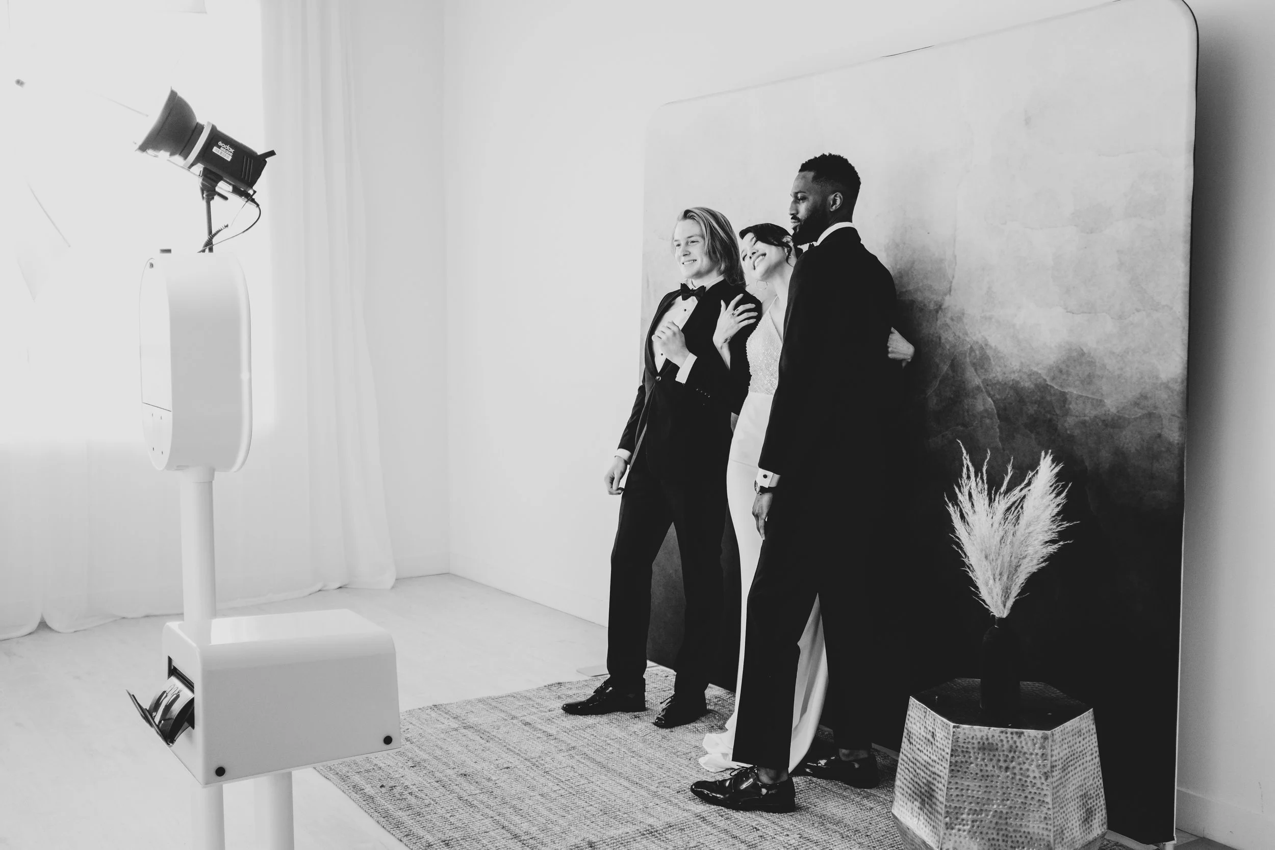 Three people posing for photos at a photo booth, with one woman in a white dress and two men in black tuxedos, standing in front of a decorative backdrop with a plant and textured wall.