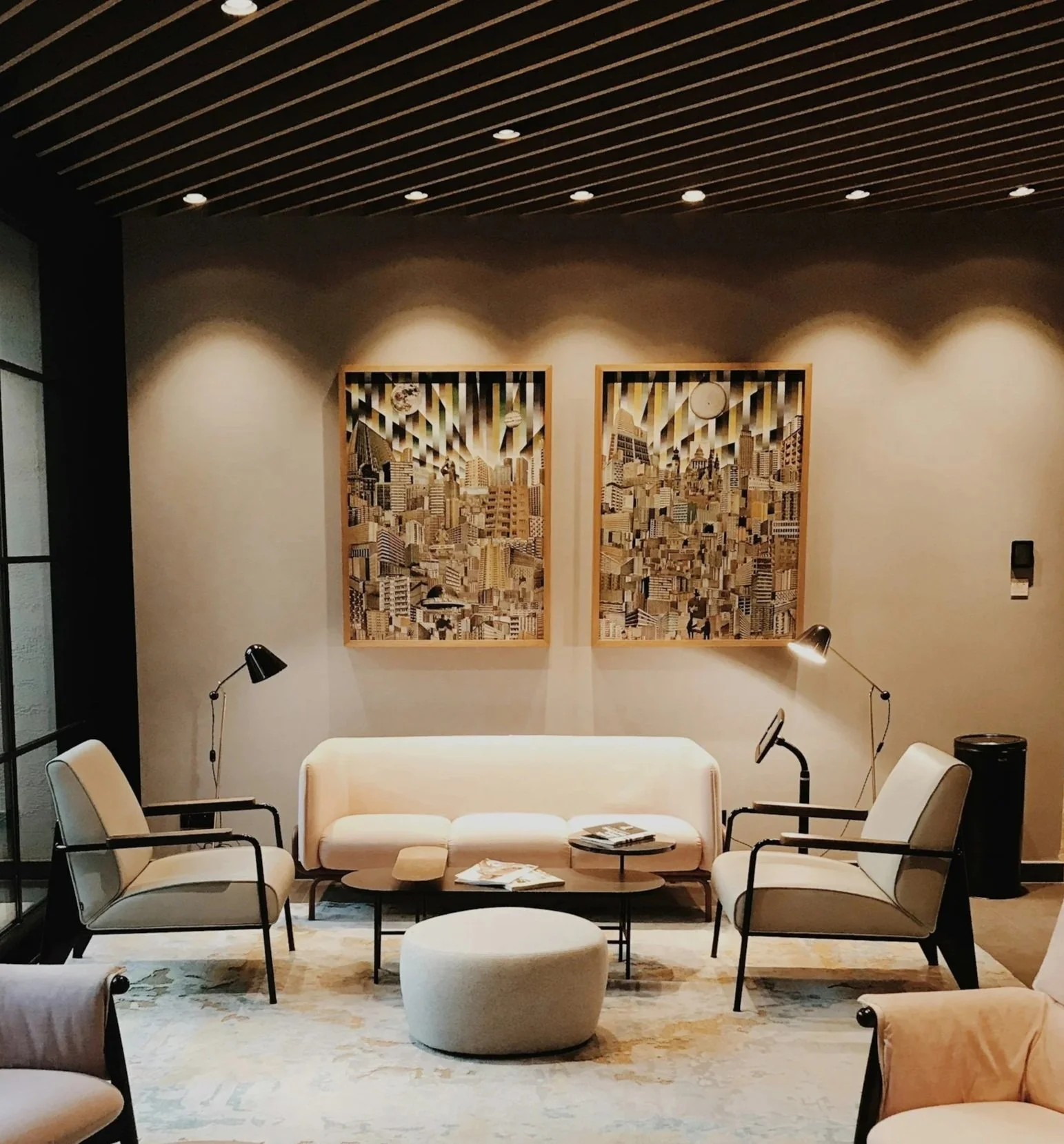 A modern living room with a beige sofa, two white armchairs with black metal arms, a round cream ottoman, and a glass-topped coffee table with reading materials. Two black adjustable reading lamps are directed towards the sitting area. Two framed cityscape artworks made of variously colored wood are hung on a beige wall. The ceiling has wooden slats with embedded lights, and there is a patterned rug on the floor.
