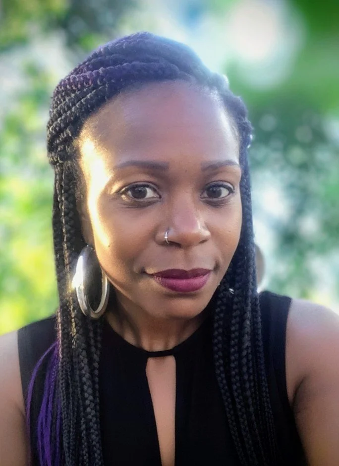 A woman with braided hair, hoop earrings, and a nose ring takes a selfie outdoors with green foliage in the background.