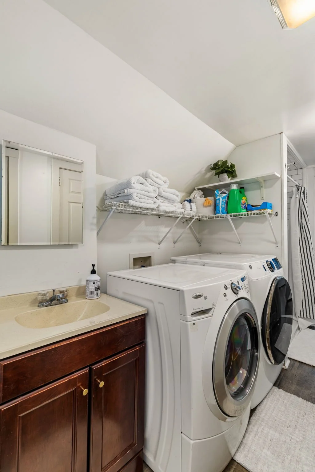 A laundry room with a washing machine and dryer against the wall, a shelf with folded towels, cleaning supplies, and a potted plant, a small sink with soap, and a mirror on the wall.