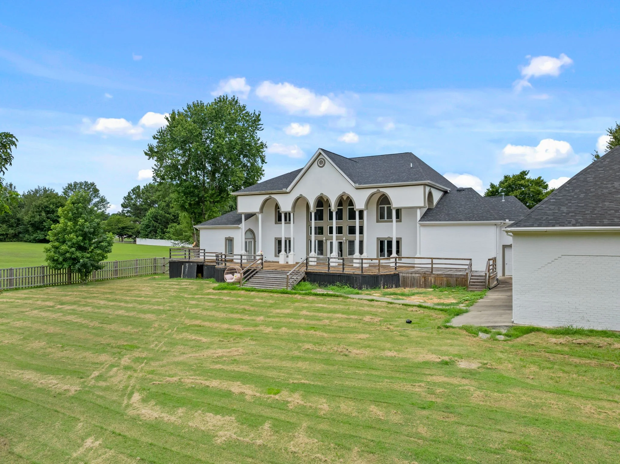 Large white house with a dark gray roof and arched windows, surrounded by greenery and trees under a blue sky with white clouds.