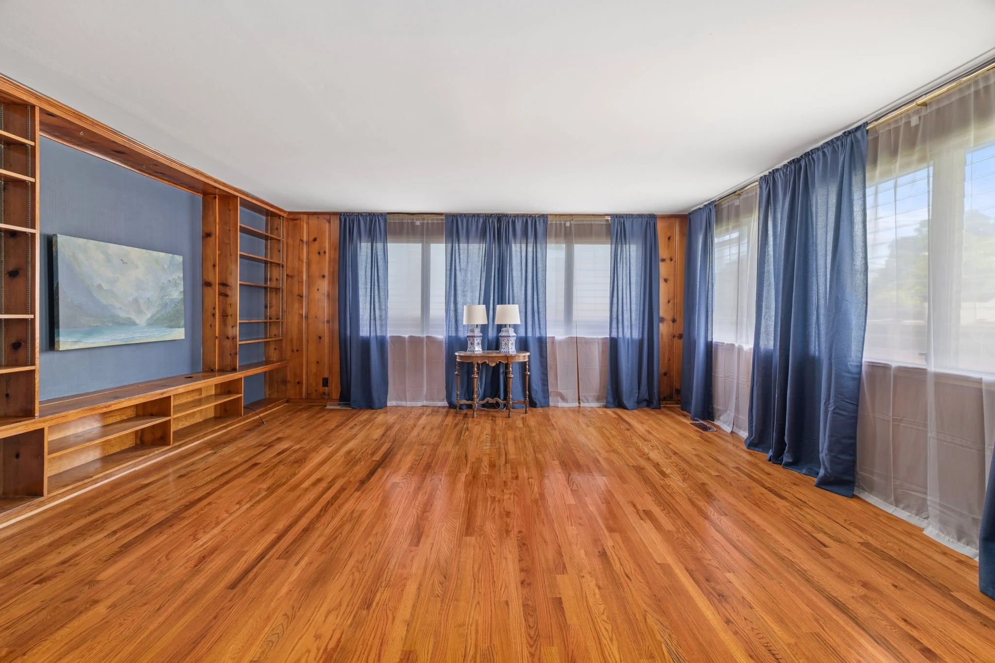 Living room with hardwood floors, blue curtains, wooden wall paneling, a small wooden table with two lamps, a large window, and a blue wall-mounted shelf with a painting.