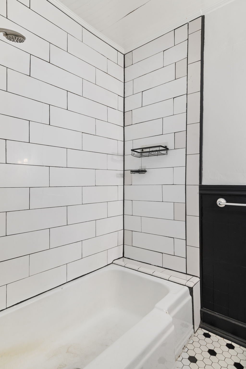 Bathroom shower with white subway tile walls, black trim, a built-in soap dish, and a showerhead, with a partial view of black bathroom cabinetry and hexagonal black and white tiled floor.