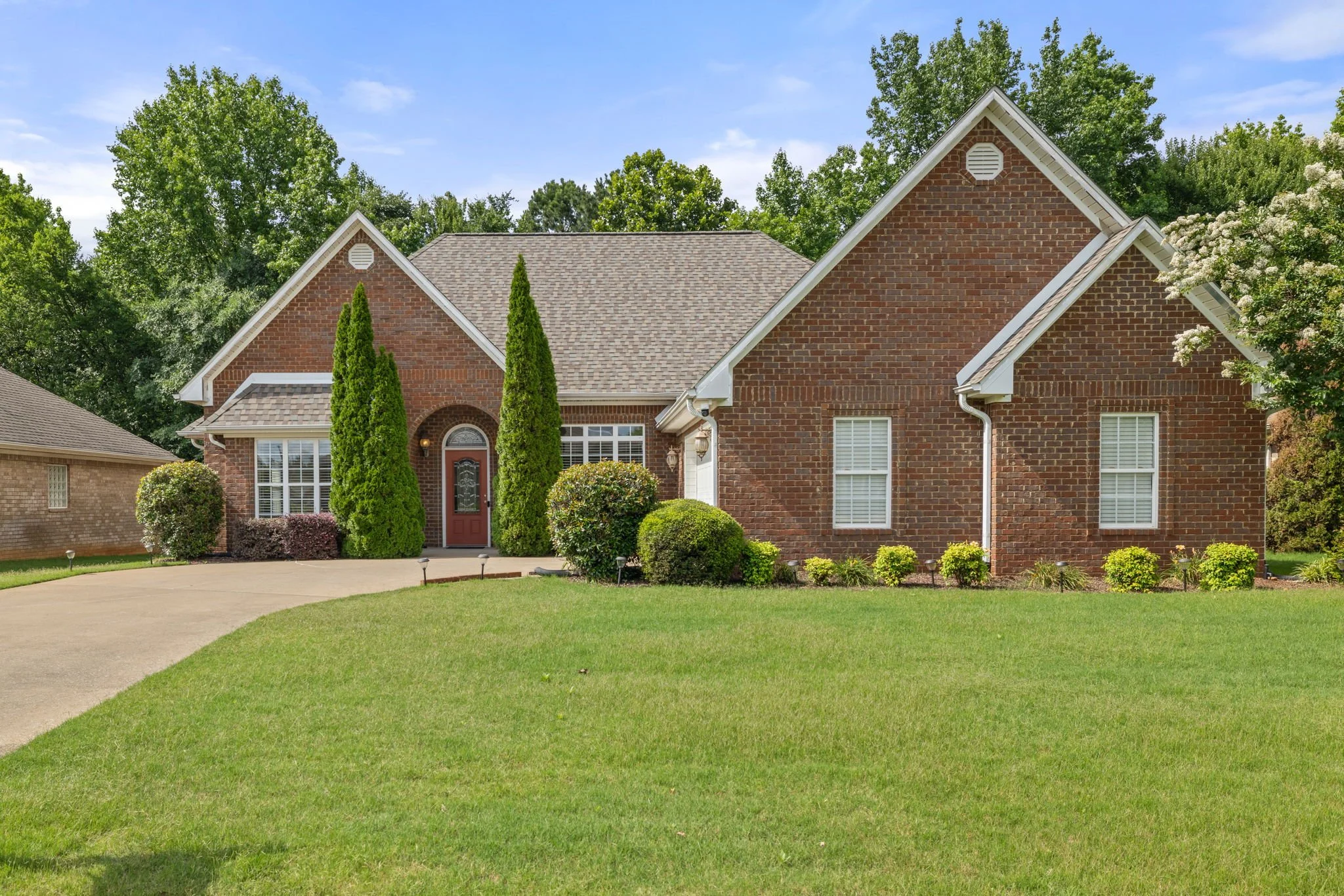 A red brick house with a gabled roof, surrounded by a green lawn and bushes, with tall trees and a partly cloudy sky in the background.