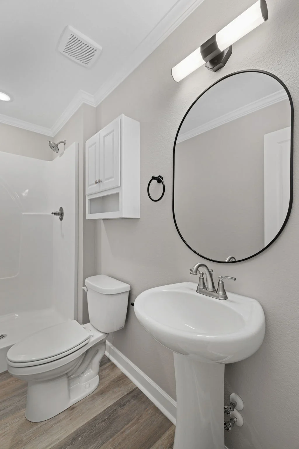A small bathroom with a walk-in shower, a white toilet, a pedestal sink, an oval mirror, a wall-mounted black towel ring, and a white wall cabinet. The bathroom has light-colored walls and wood-like flooring.