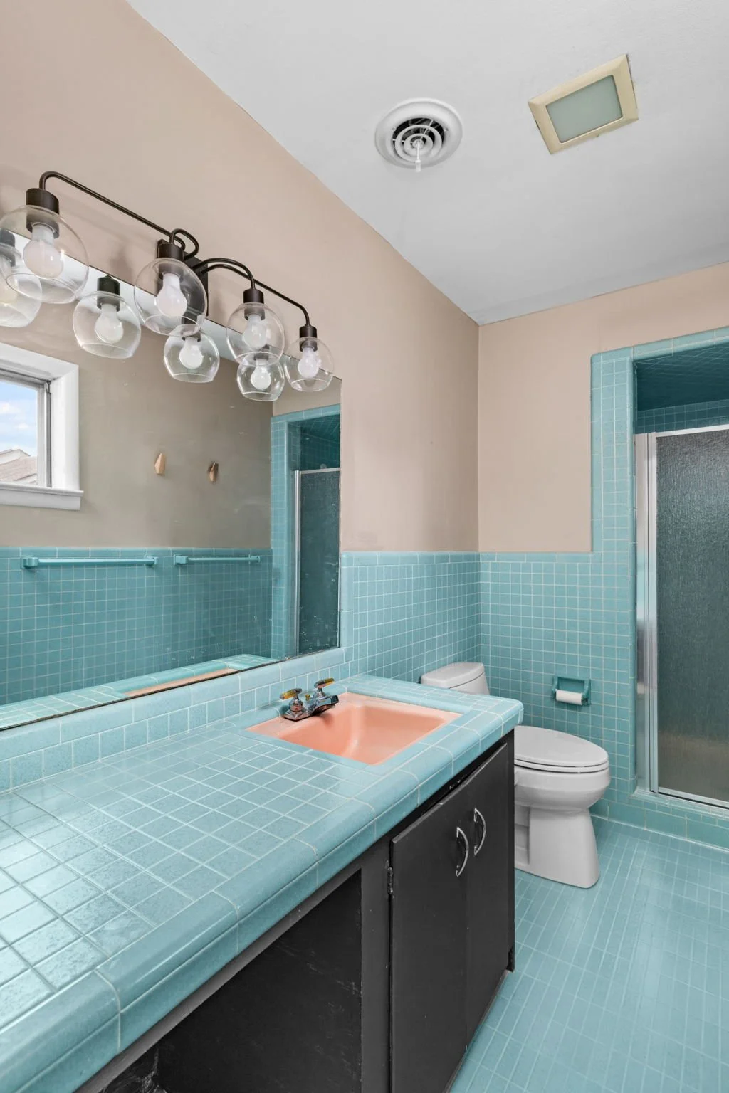 Bathroom with light blue tiled walls and floor, peach-colored sink, black vanity, toilet, shower with glass door, small window, large mirror, and ceiling light fixture.