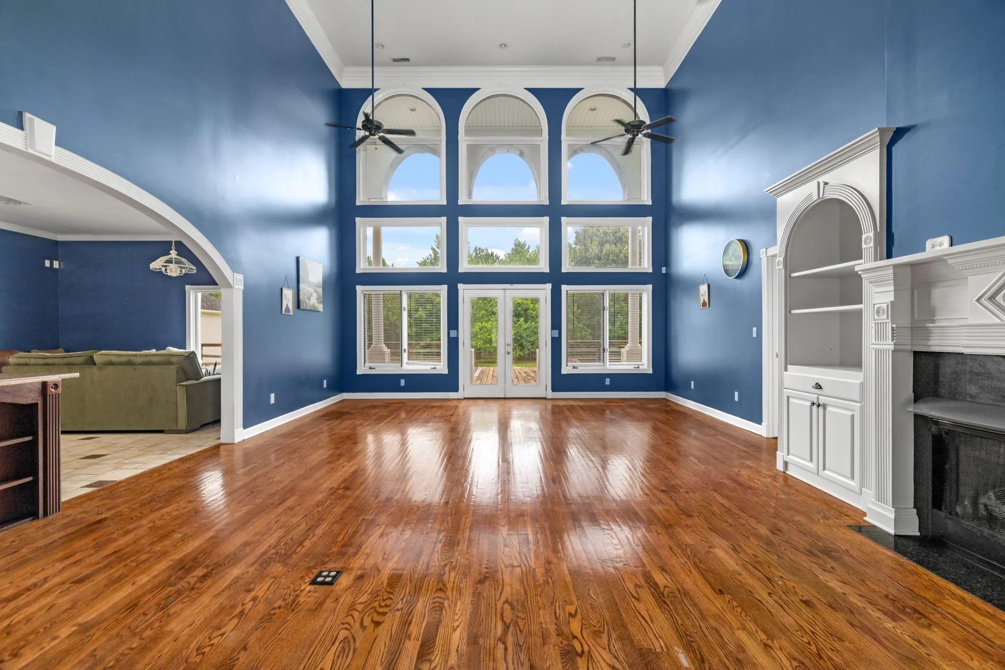 Spacious living room with large blue walls, tall windows, hardwood floors, and a fireplace with white mantel. Ceiling fans are visible, and patio doors lead to an outdoor deck.