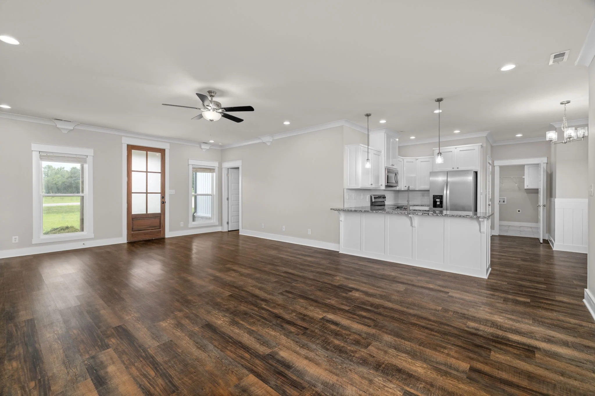 Open-concept living space with hardwood floors, white kitchen cabinets, stainless steel appliances, a ceiling fan, and large windows.