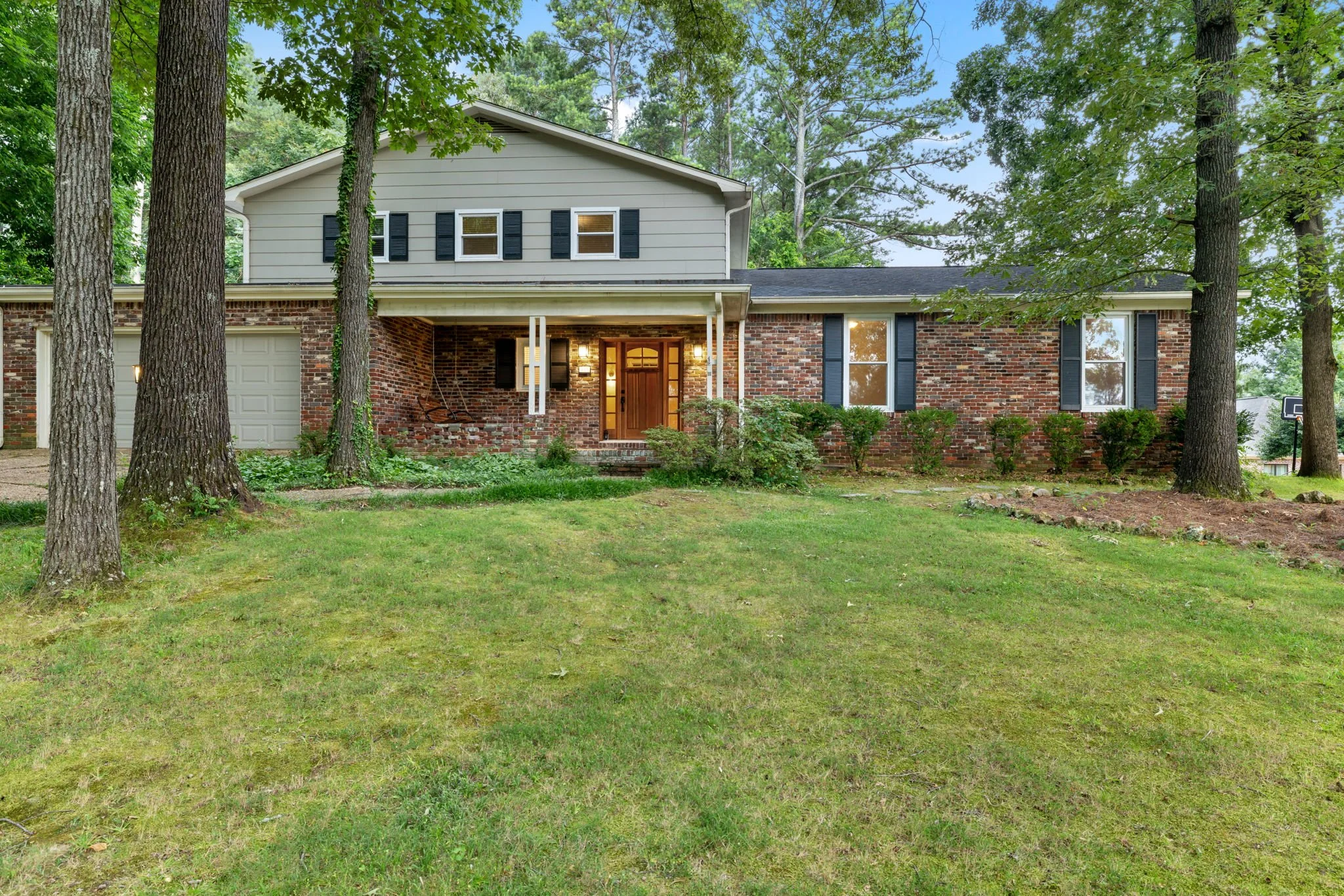 Front view of a house with a brick lower level and a beige upper level, surrounded by tall trees and a grassy lawn.