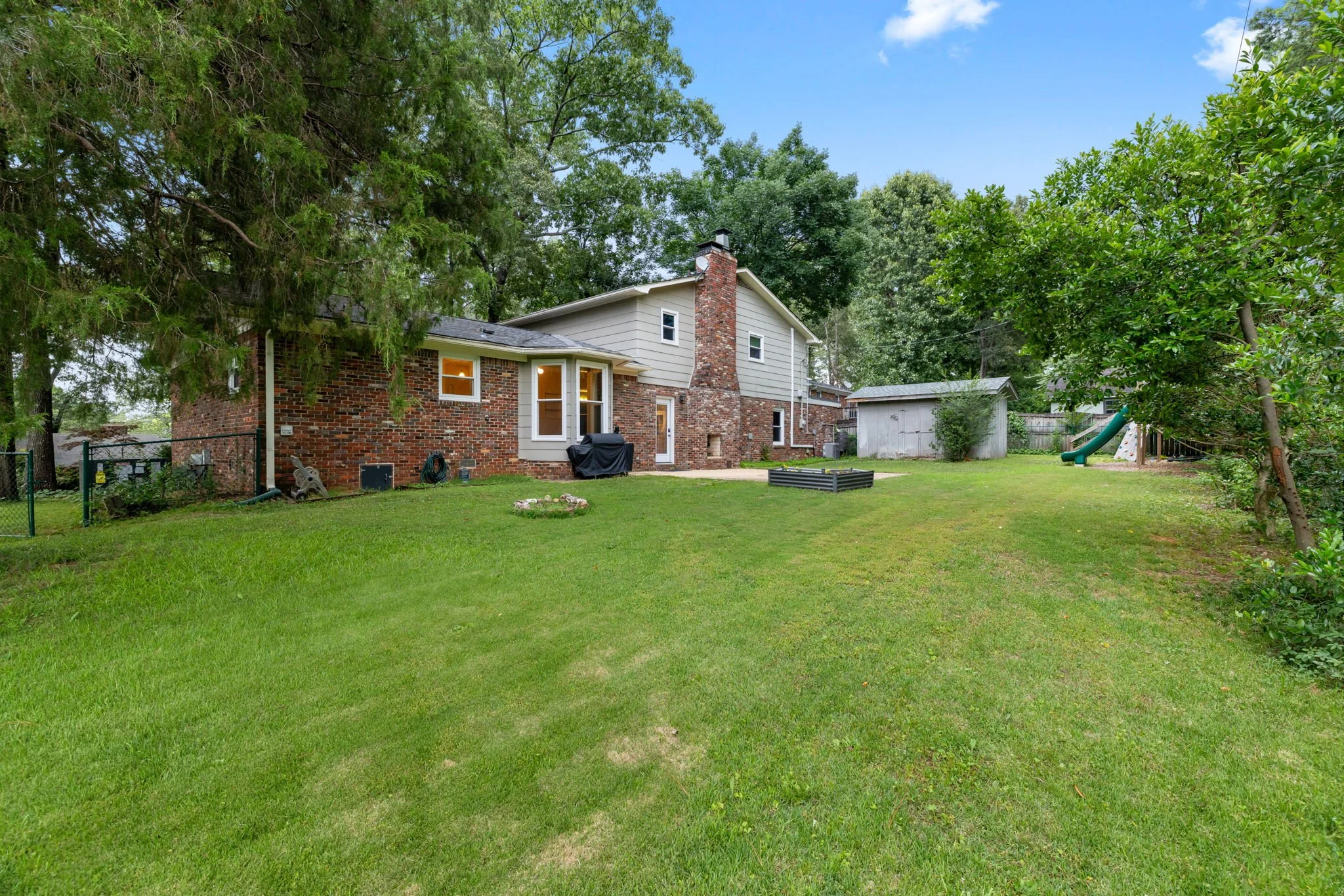 Backyard with a green lawn, large trees, a brick house with a chimney, a small storage shed, and a playground slide.