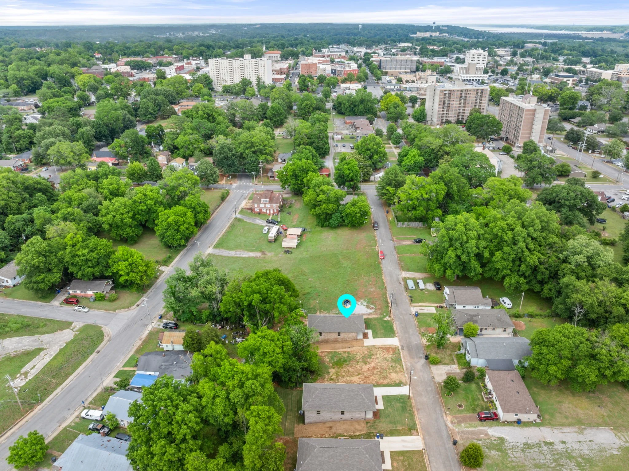 An aerial view of a city with a mix of residential houses, tall apartment buildings, and brick commercial buildings, surrounded by green trees and streets.