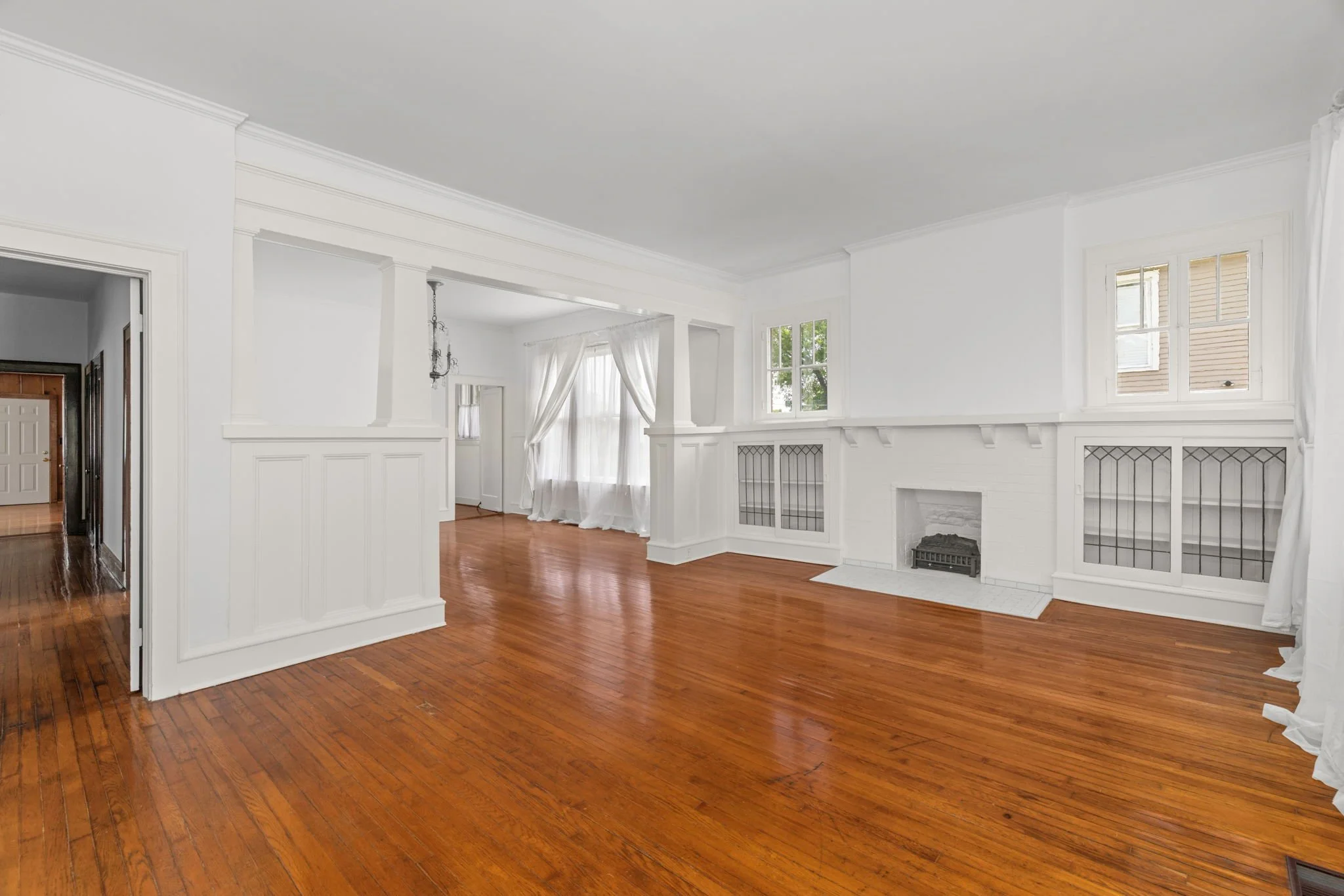 Bright living room with white walls and large windows, hardwood floors, and a white fireplace.