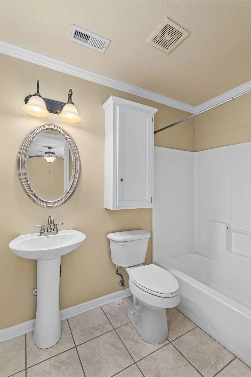 Bathroom with a pedestal sink, oval mirror, vanity light, toilet, white shower stall, beige walls, tiled floor, and ceiling vent.