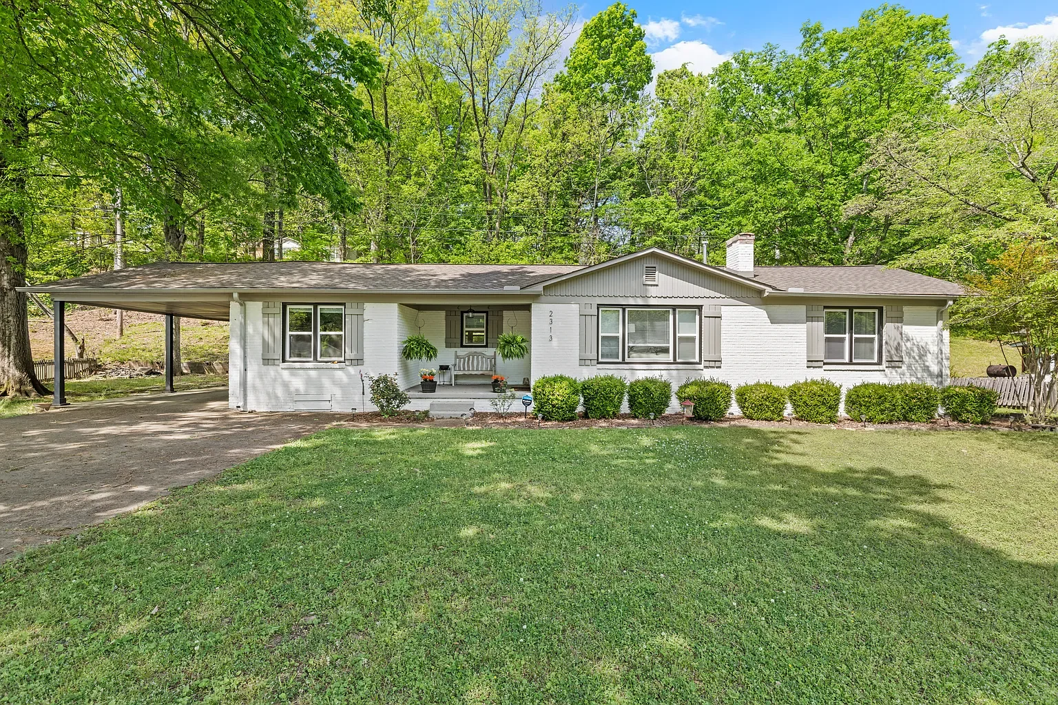 A single-story white house with grey shutters, a front porch with two hanging plants, and a small green lawn surrounded by trees.
