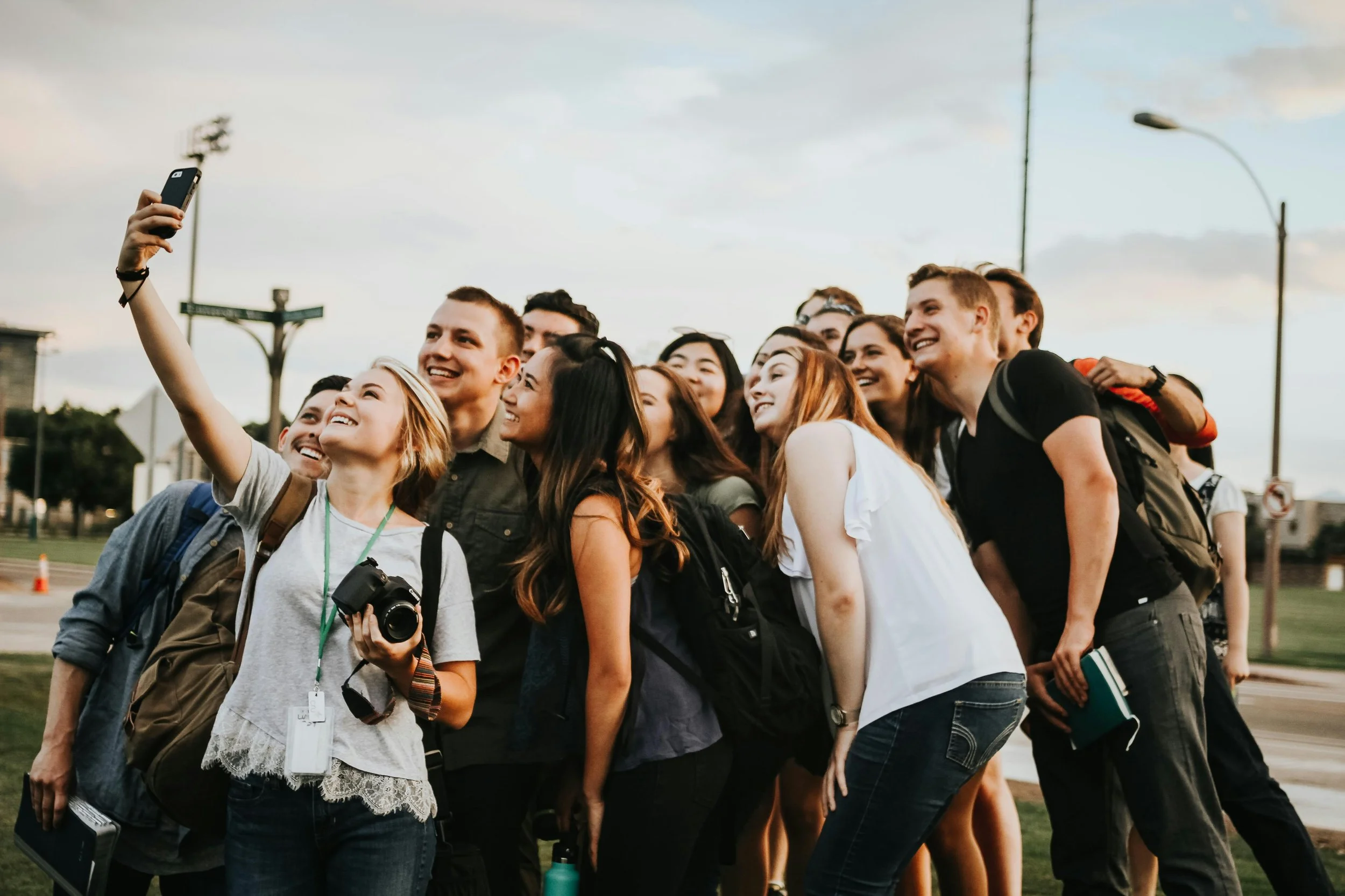 A group of young people taking a selfie outdoors during daytime. They are smiling and standing close together, with some holding cameras and notebooks.