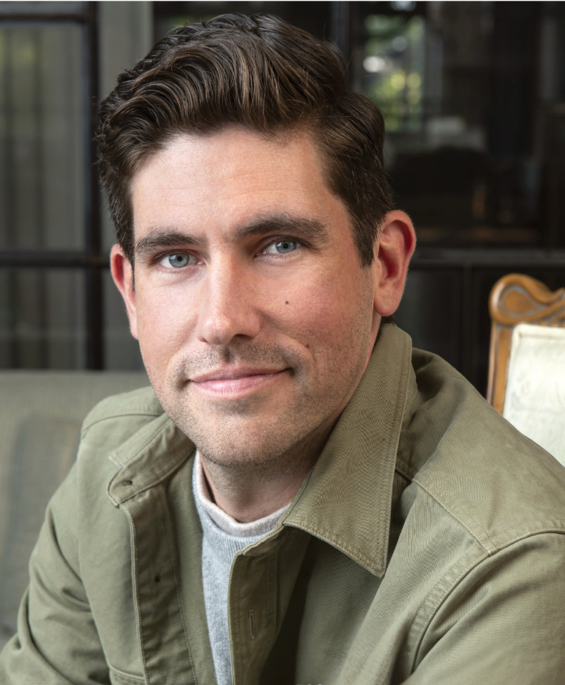 A man with short brown hair, blue eyes, and light skin, sitting indoors in a casual setting, wearing a green jacket over a gray shirt, smiling softly at the camera.