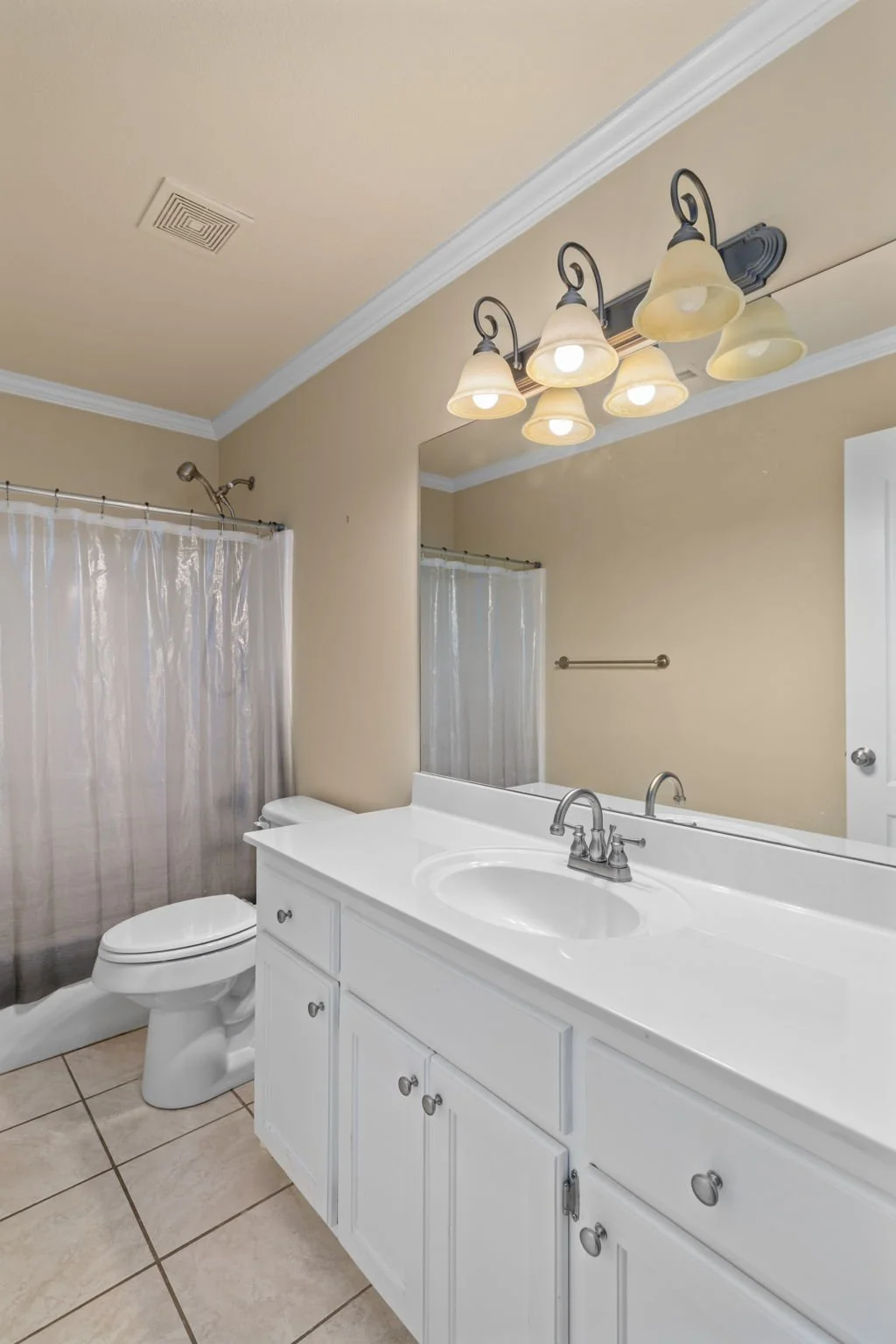 Bathroom with a white vanity, sink, large mirror, beige walls, four-light fixture, toilet, and shower with a gray curtain. Tiled floor and crown molding.