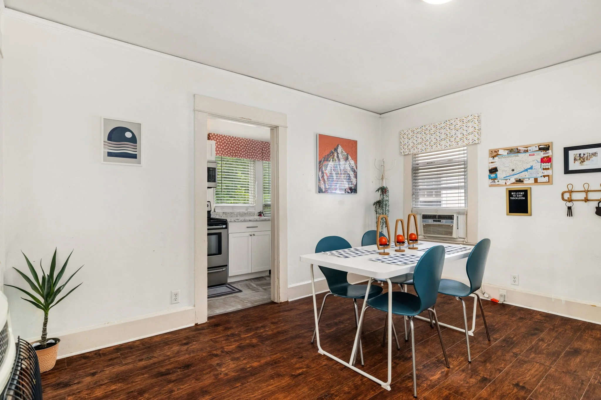 A living room with a wooden floor and white walls, featuring a dining table with four teal chairs, a wall decorated with paintings and a corkboard, and a view into the kitchen with a window, oven, and microwave.