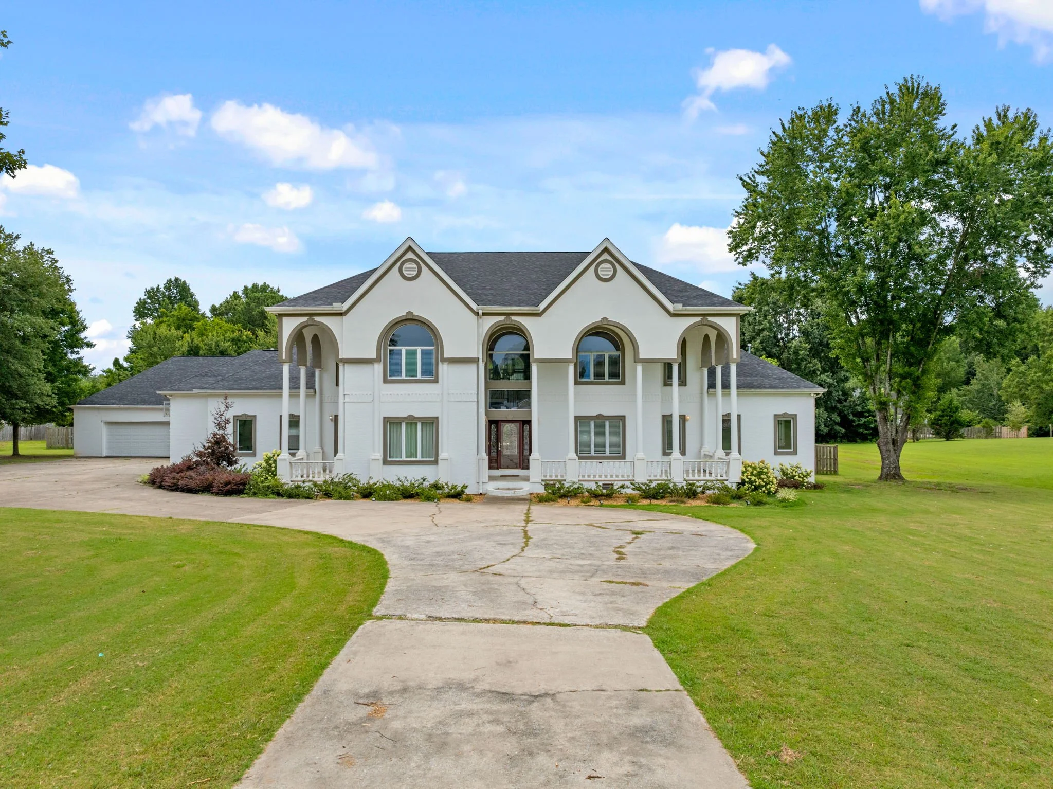 Front view of a large white house with a driveway, surrounded by green grass, trees, and blue sky with white clouds.
