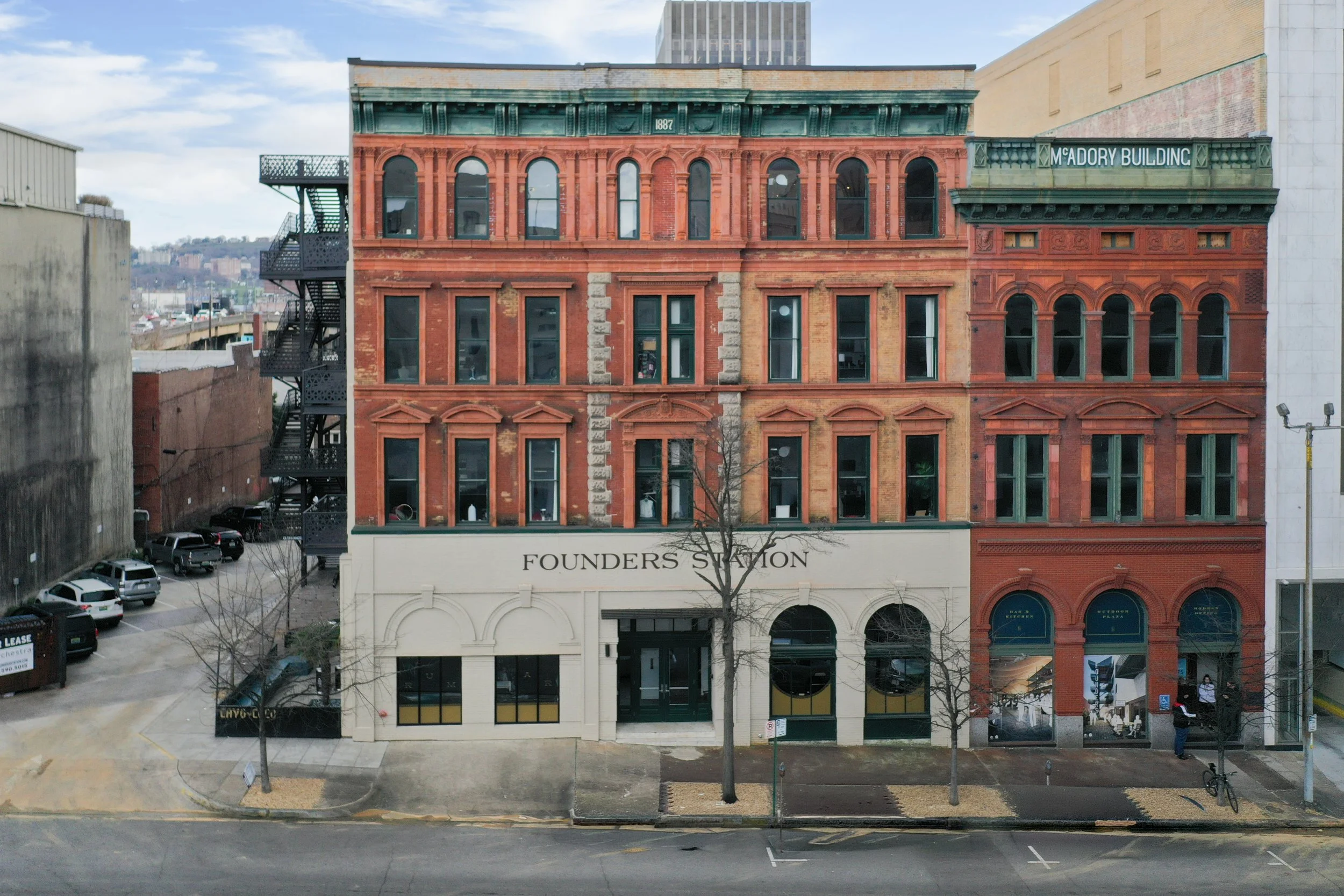 A four-story red brick building with architectural details atop a white ground-floor storefront, labeled 'Founders Station' in black text. The upper floors feature large arched windows, some with visible furniture or objects inside. Adjacent to the building are parking lots and trees with barren branches; in the background, there are more city buildings and a partly cloudy sky.