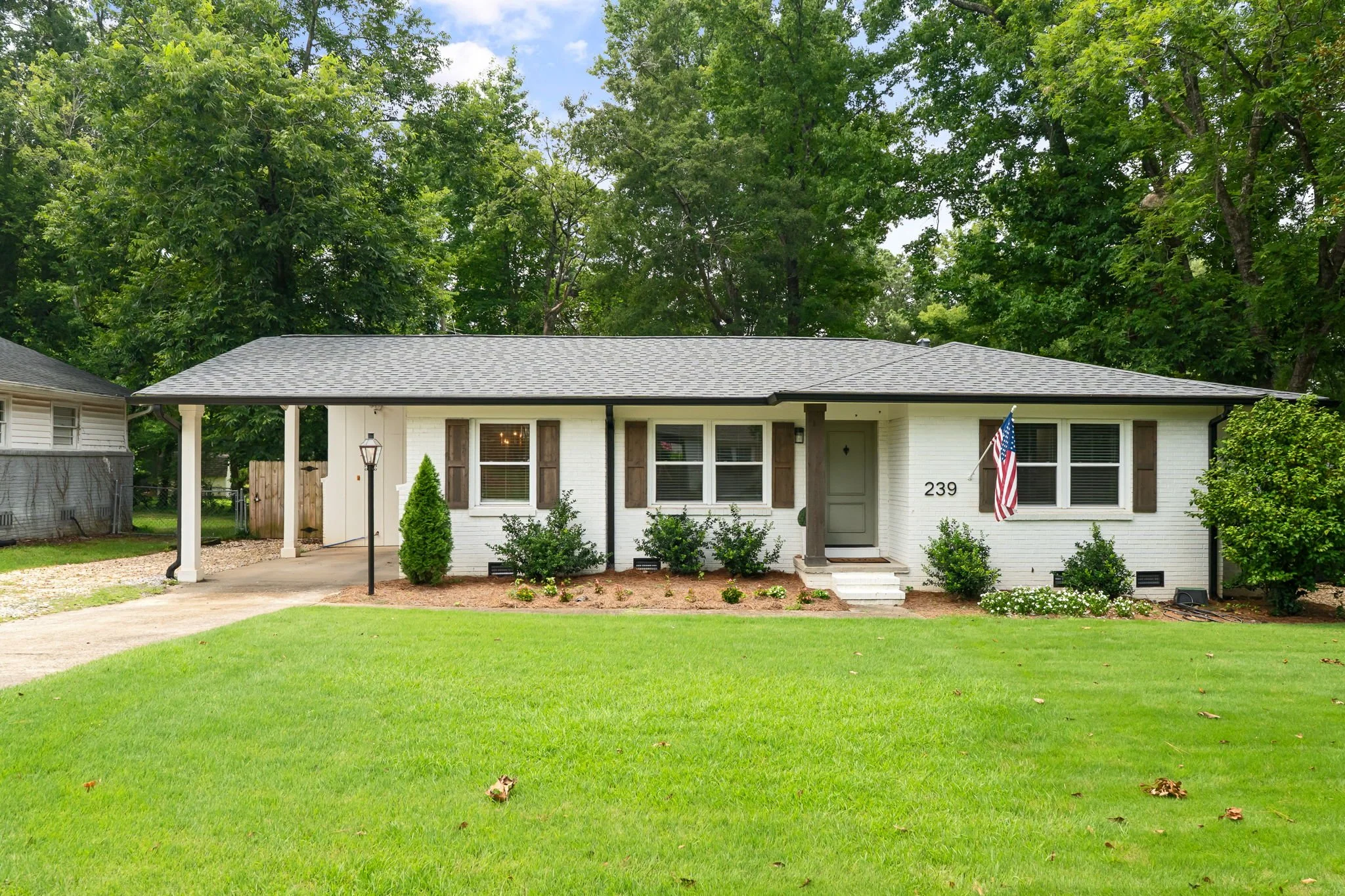Front view of a white house with brown shutters and an American flag hanging outside. The house has a gray shingle roof and is surrounded by green grass, small bushes, and tall trees in the background.