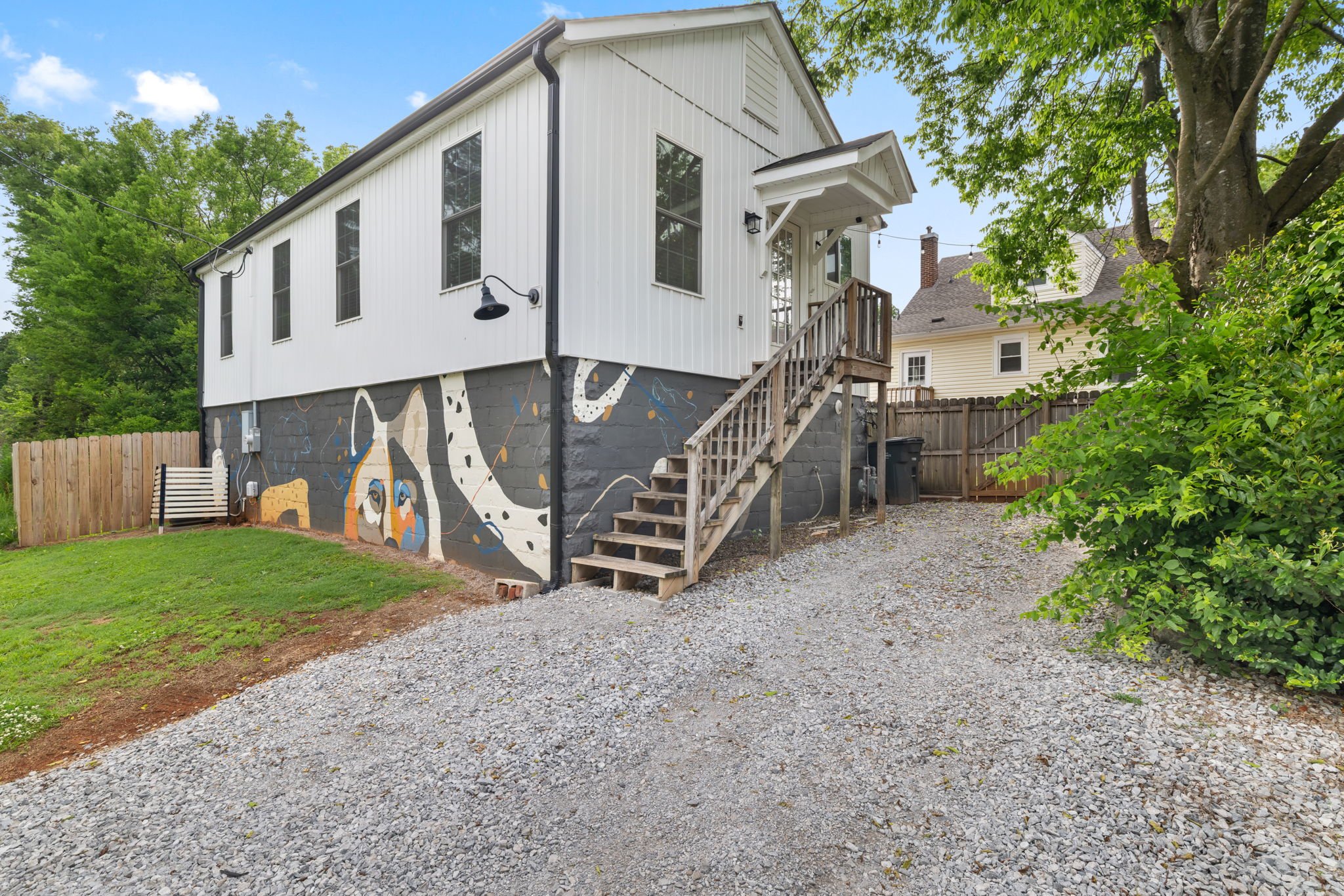 Backyard of a house with painted mural on the side wall, depicting a Dalmatian dog with blue and beige accents. Wooden stairs lead to a small door, and there is a black wall-mounted light fixture. The yard has a grassy area and a gravel pathway, surrounded by trees and a wooden fence.