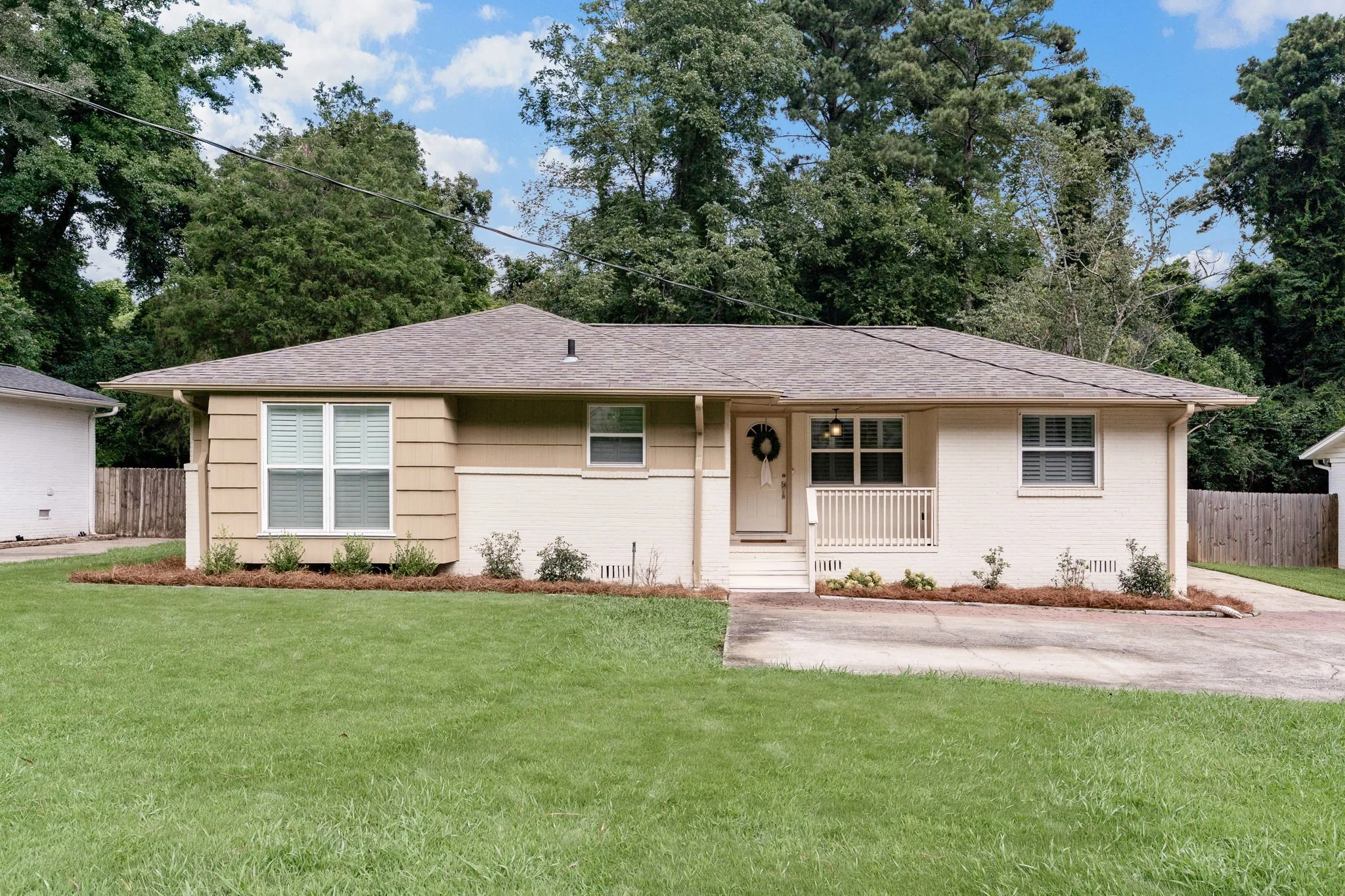 Single-story house with a beige and white exterior, front porch with railing, surrounded by a green lawn, and large trees in the background under partly cloudy sky.