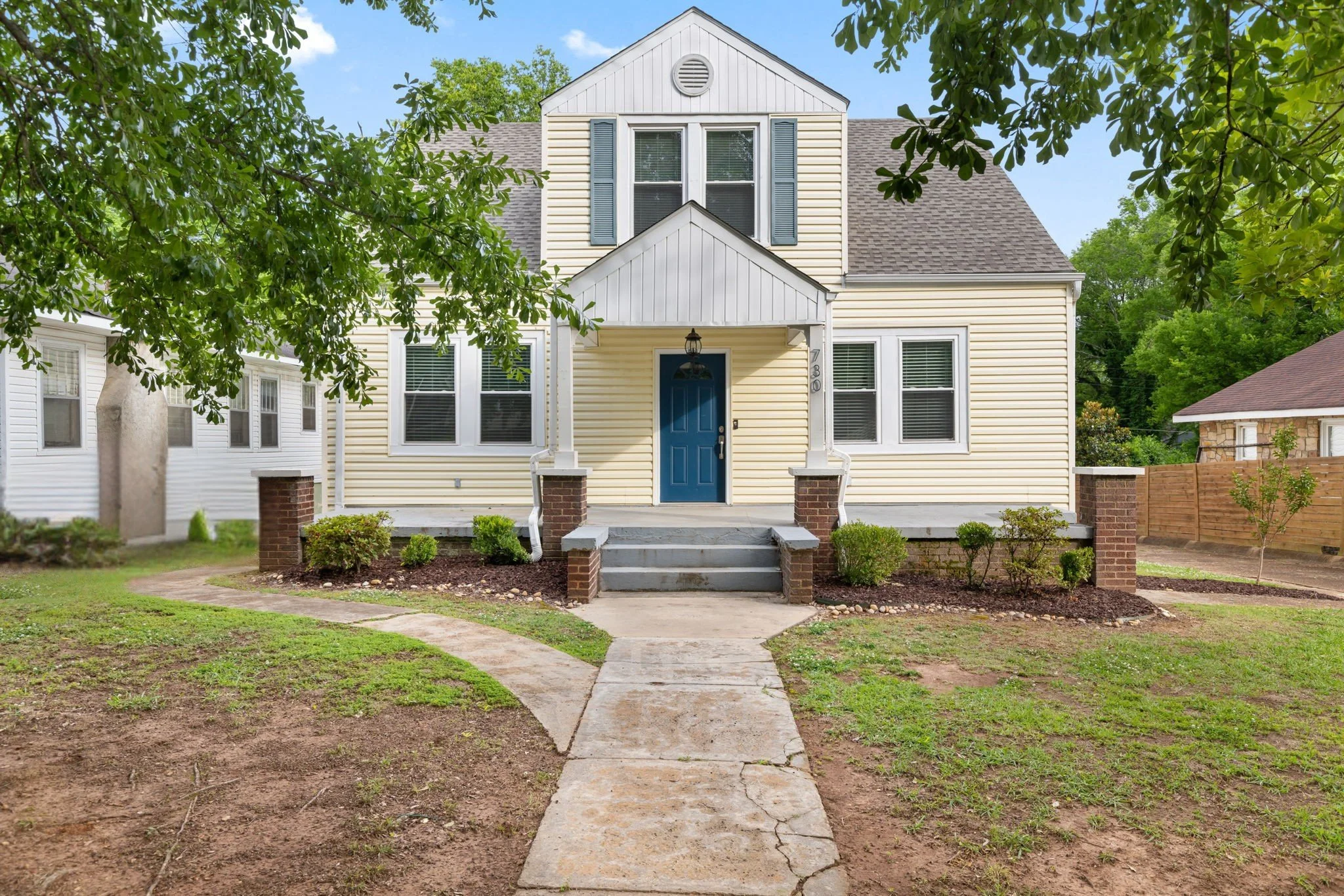 Front view of a light yellow, two-story house with blue front door, surrounded by trees and a garden with a concrete pathway.