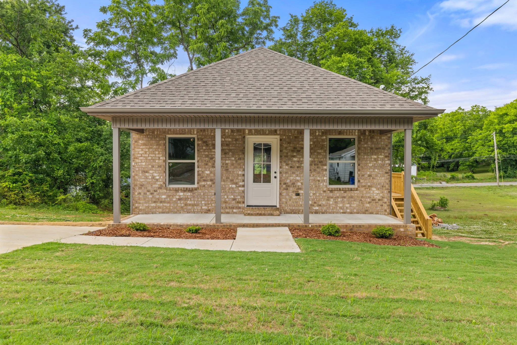 A house with brick walls, a gabled roof, two windows, a front door, a small porch, and stairs on the right side, surrounded by green trees and a grassy yard.