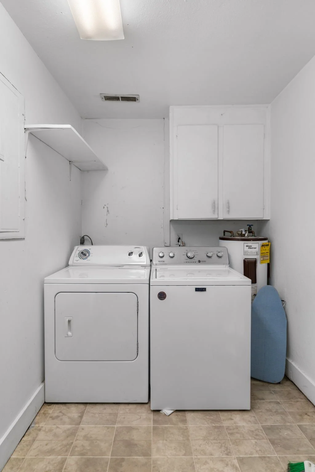 A laundry room with a white washing machine on the left and a white dryer on the right. There is a small cabinet mounted on the wall above the dryer, and a water heater in the corner. An ironing board leans against the wall next to the water heater. The room has beige tiled flooring and white walls.