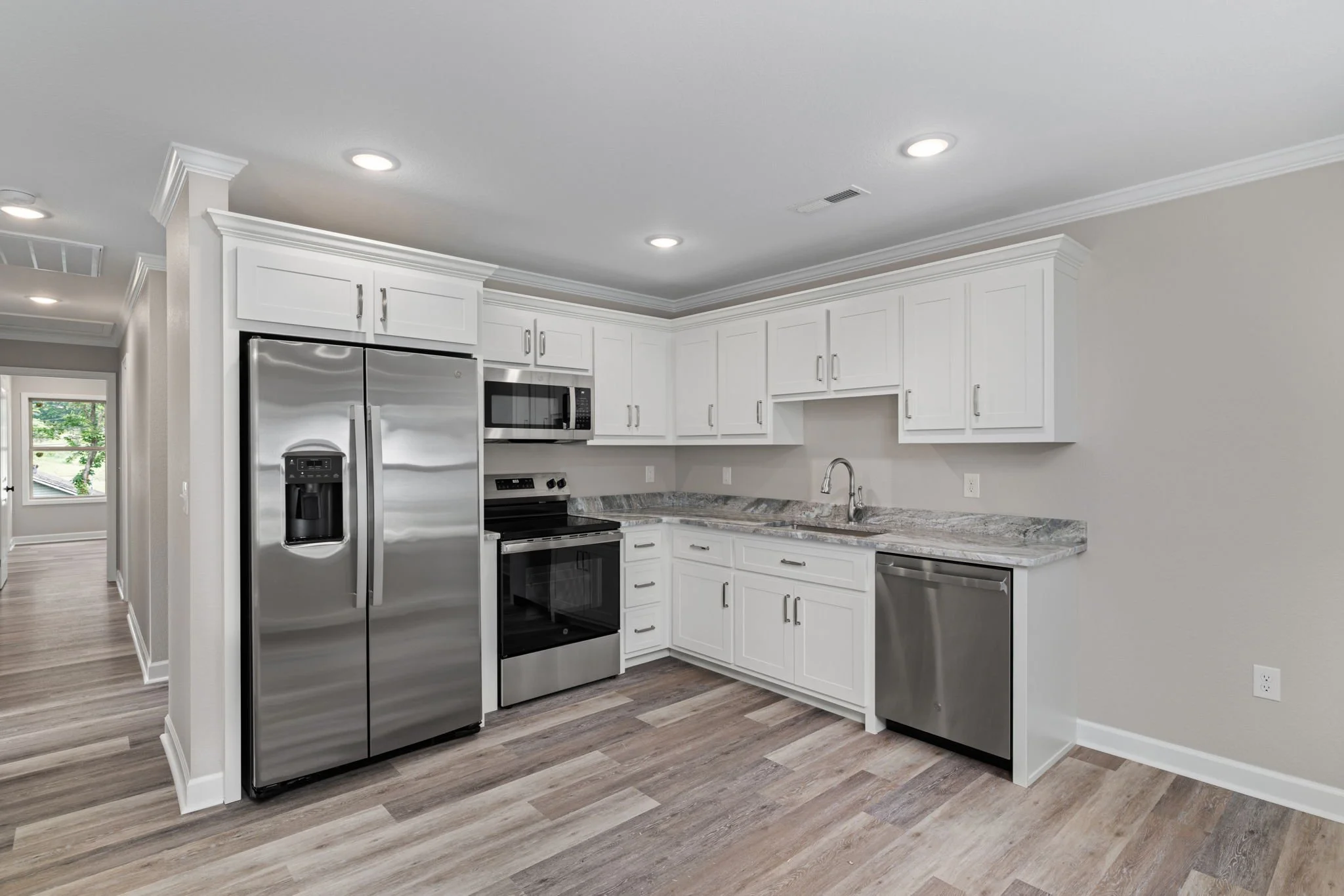 Modern kitchen with white cabinets, stainless steel refrigerator, microwave, oven, and gray marble countertops. Recessed lighting installed in the ceiling and wood flooring.