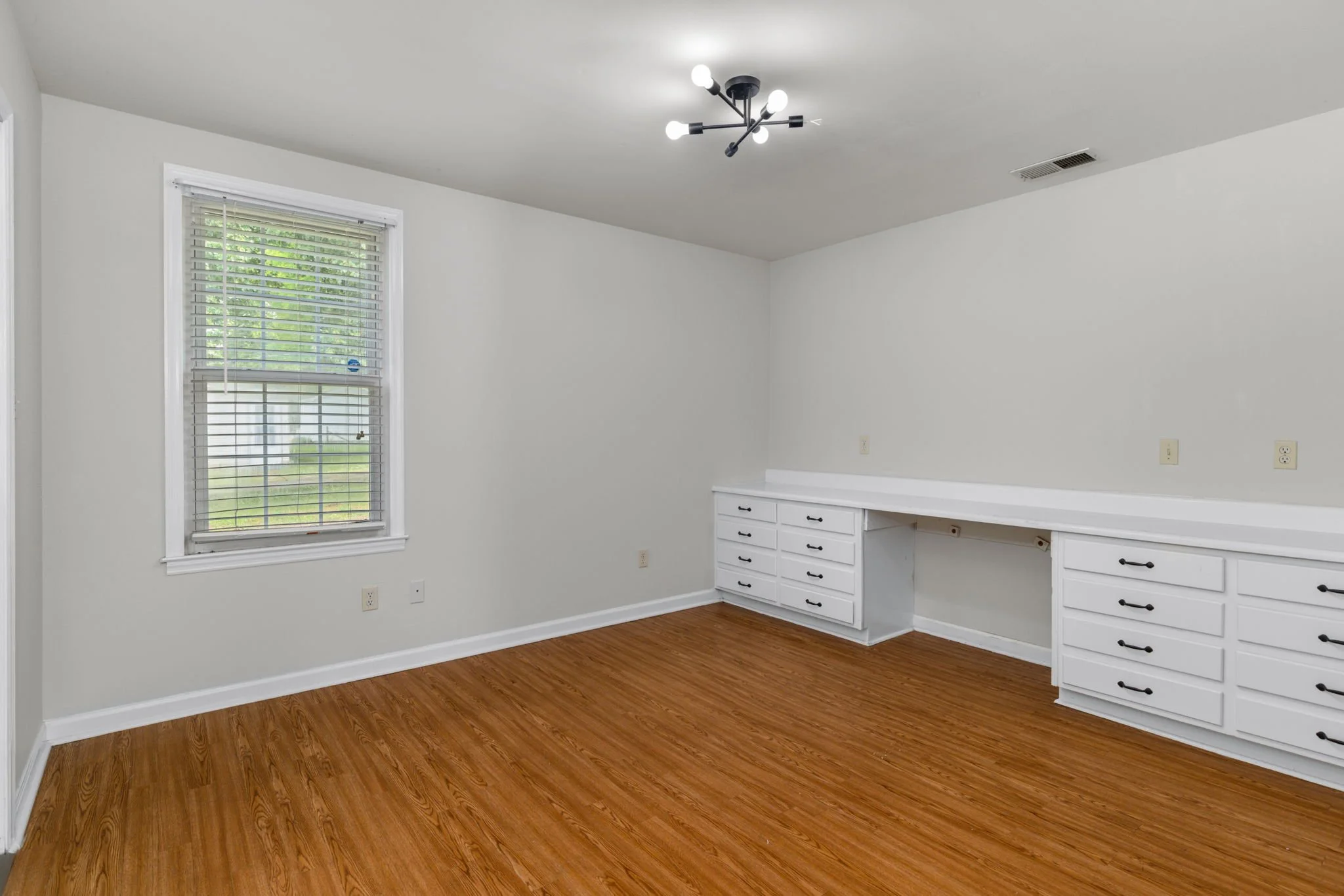 Empty room with white walls, hardwood floor, a window with blinds, a built-in white cabinet with drawers, and a modern ceiling light fixture.