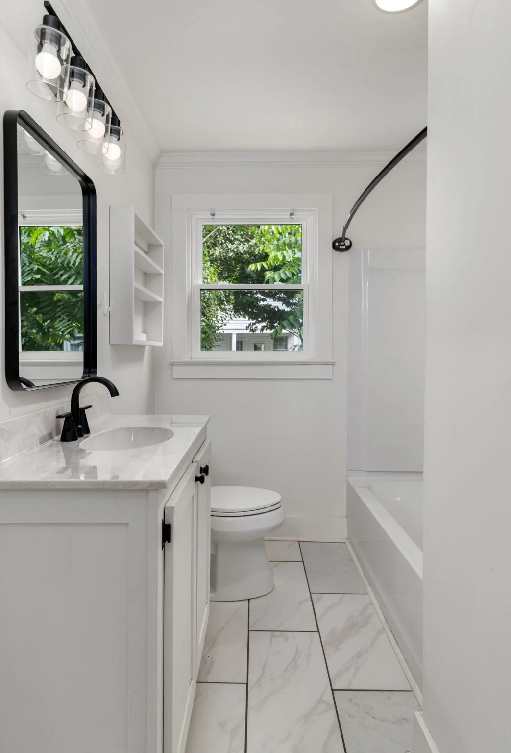 White bathroom with a vanity and sink, a toilet, a window with greenery outside, and a bathtub with a shower curtain rod.