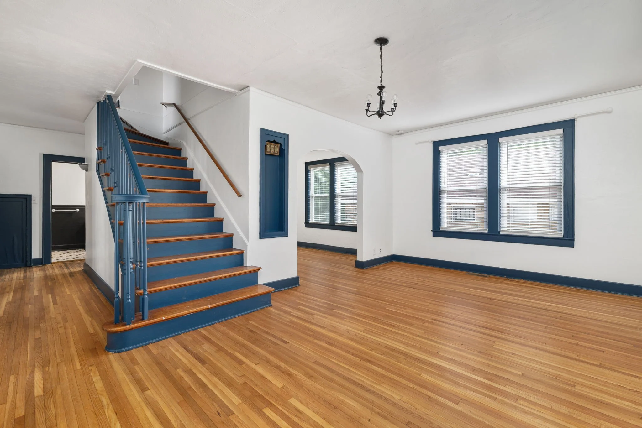 Empty, spacious living room with hardwood floors, white walls, blue trim around windows and stairs, and a black chandelier hanging from the ceiling. Ready for your furniture and personalization