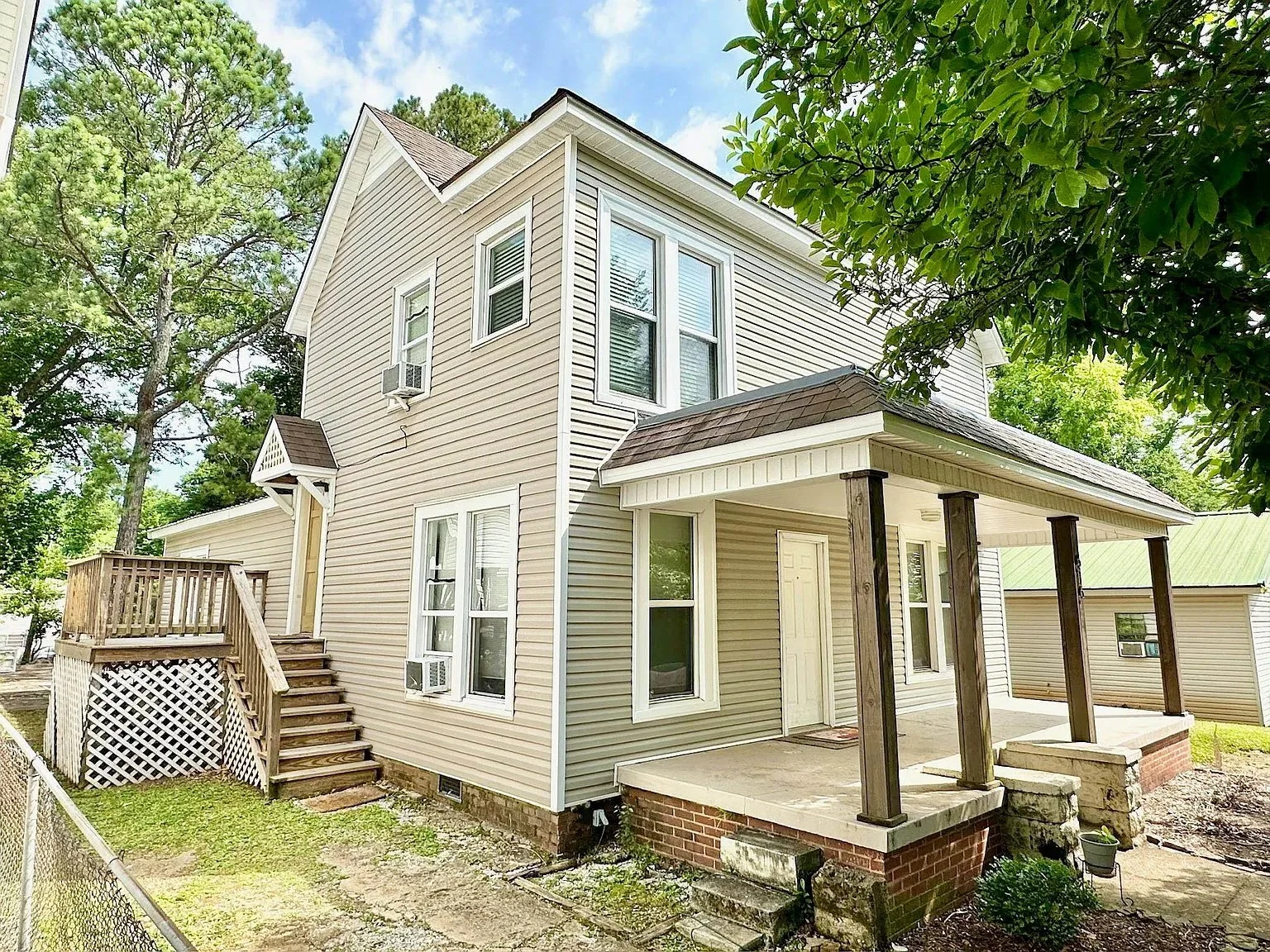 Two-story beige house with a covered front porch supported by wooden columns, multiple windows with white trim, and a staircase leading to a side door. The house is surrounded by trees and greenery.