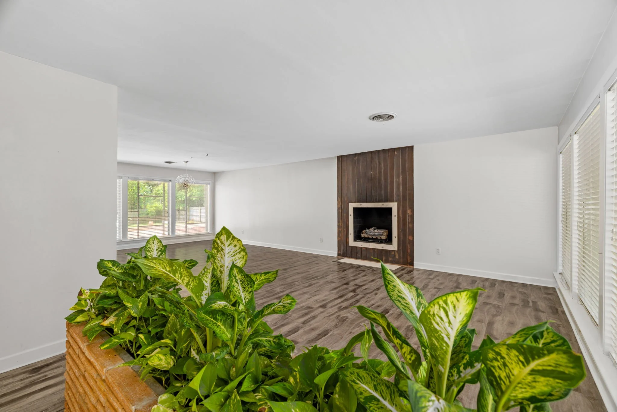 Interior view of a living room with white walls and ceiling, a fireplace with a wooden panel surround, large windows with blinds, and green plants in a brick planter in the foreground.