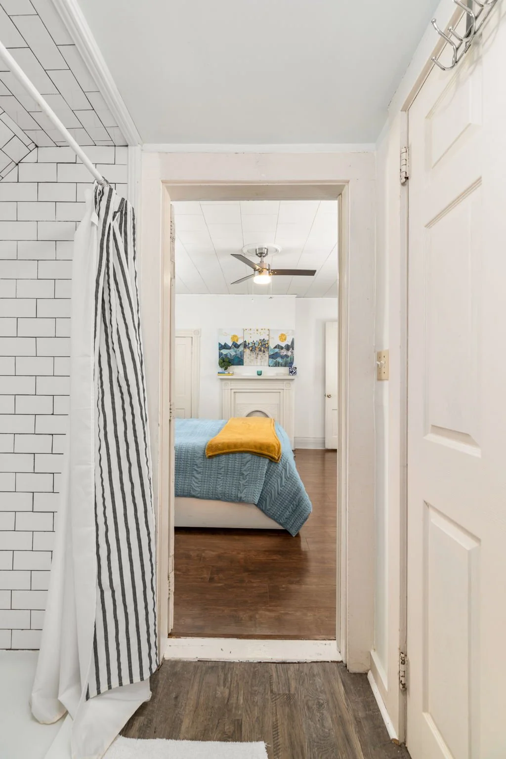 View from bathroom into bedroom with bed covered in blue quilt and yellow throw, ceiling fan, artwork above fireplace, and hardwood floors.