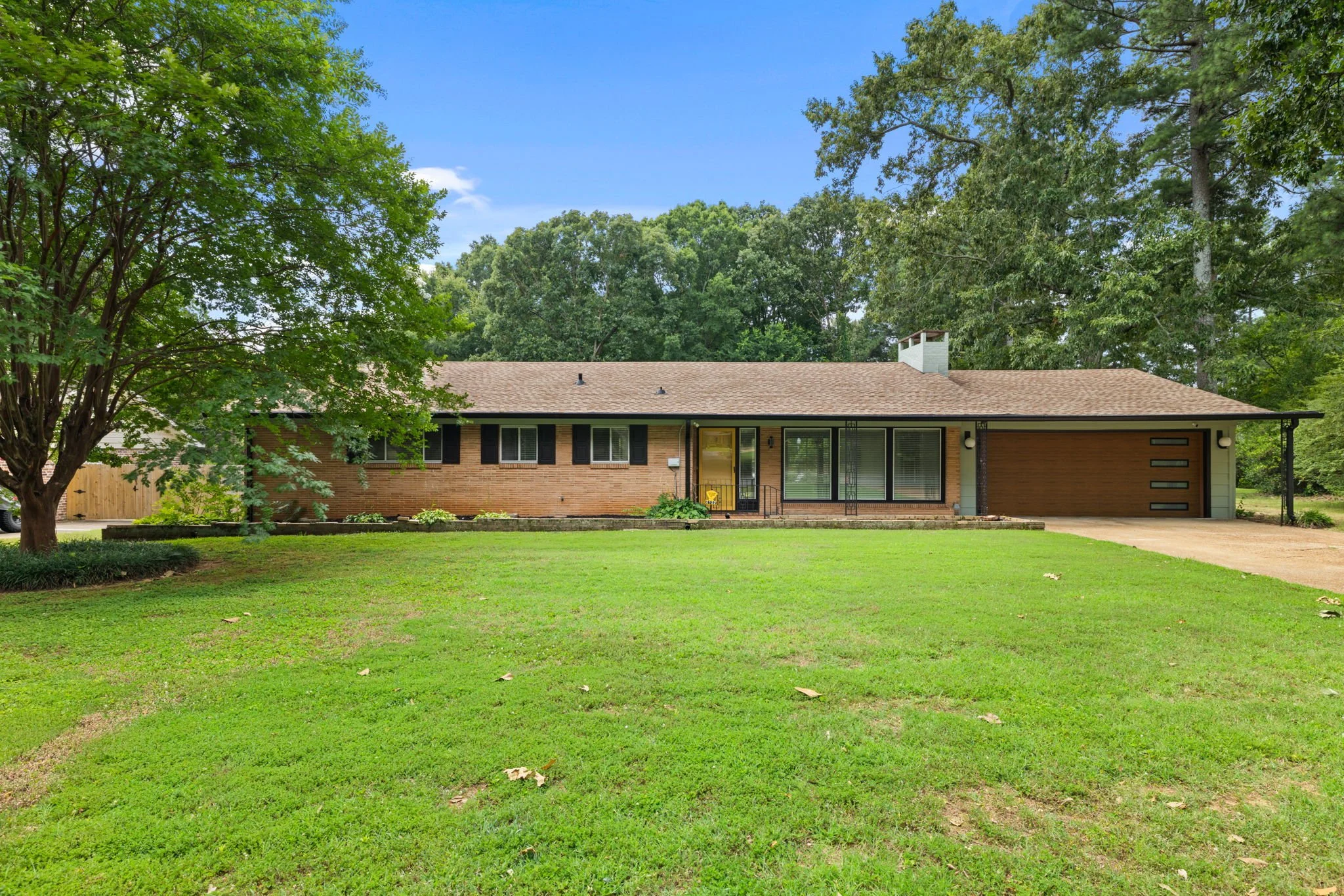 A suburban house with a brick exterior, brown garage door, and large windows, set in a lush green yard with mature trees and a bright blue sky.