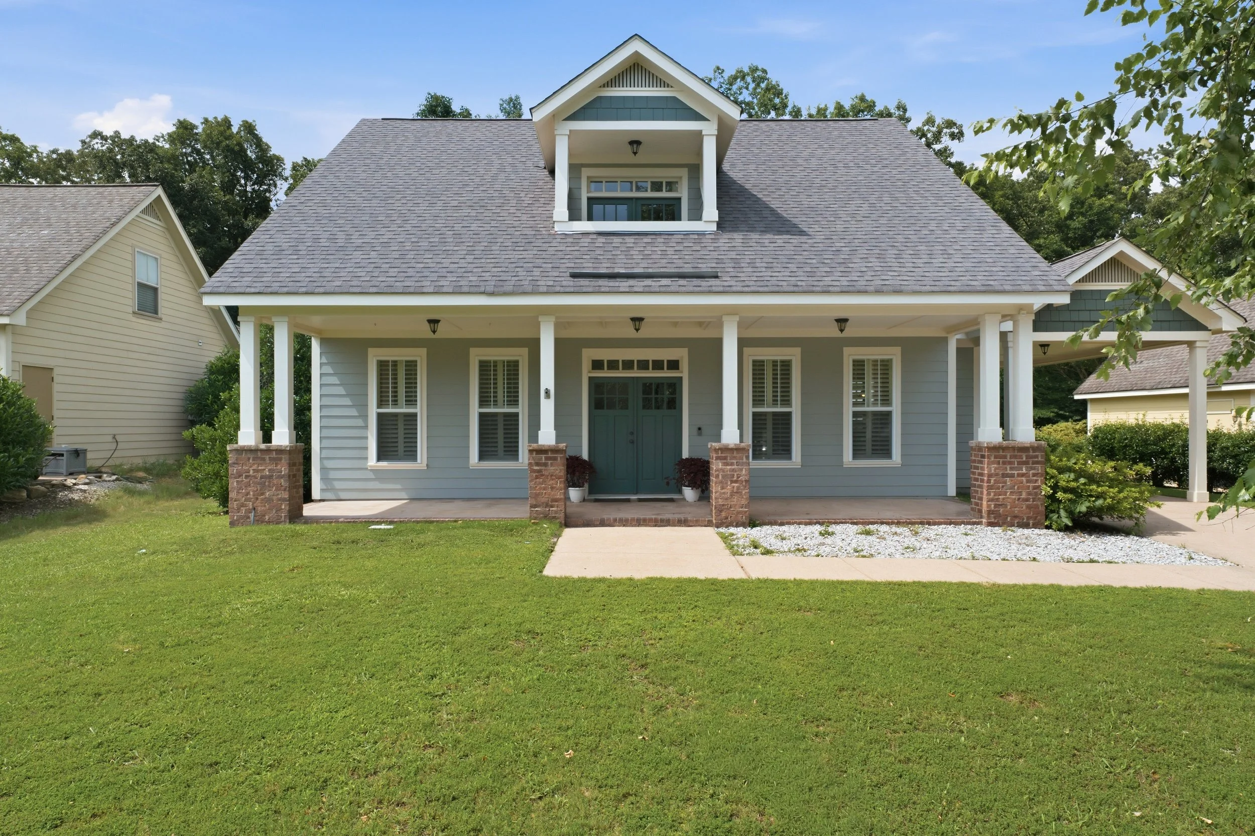 A two-story house with light blue exterior, white trim, and a front porch with brick pillars, surrounded by a green lawn and trees in the background.