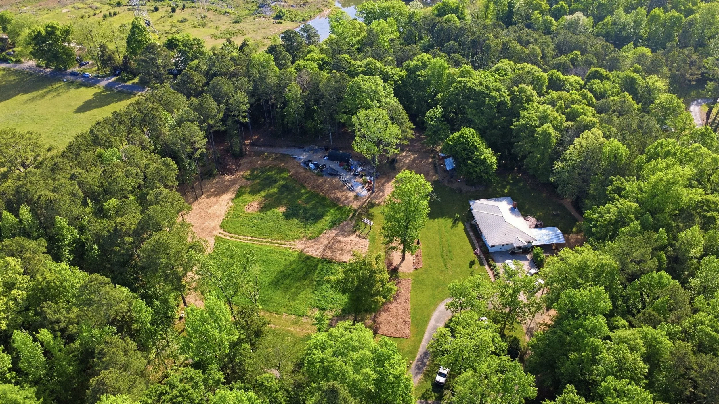 An aerial view of a backyard with a large grassy lawn, surrounding trees, a house with a deck and a driveway, and a wooded area in the background. There is a construction or installation site with vehicles and equipment near the edge of the wooded area.